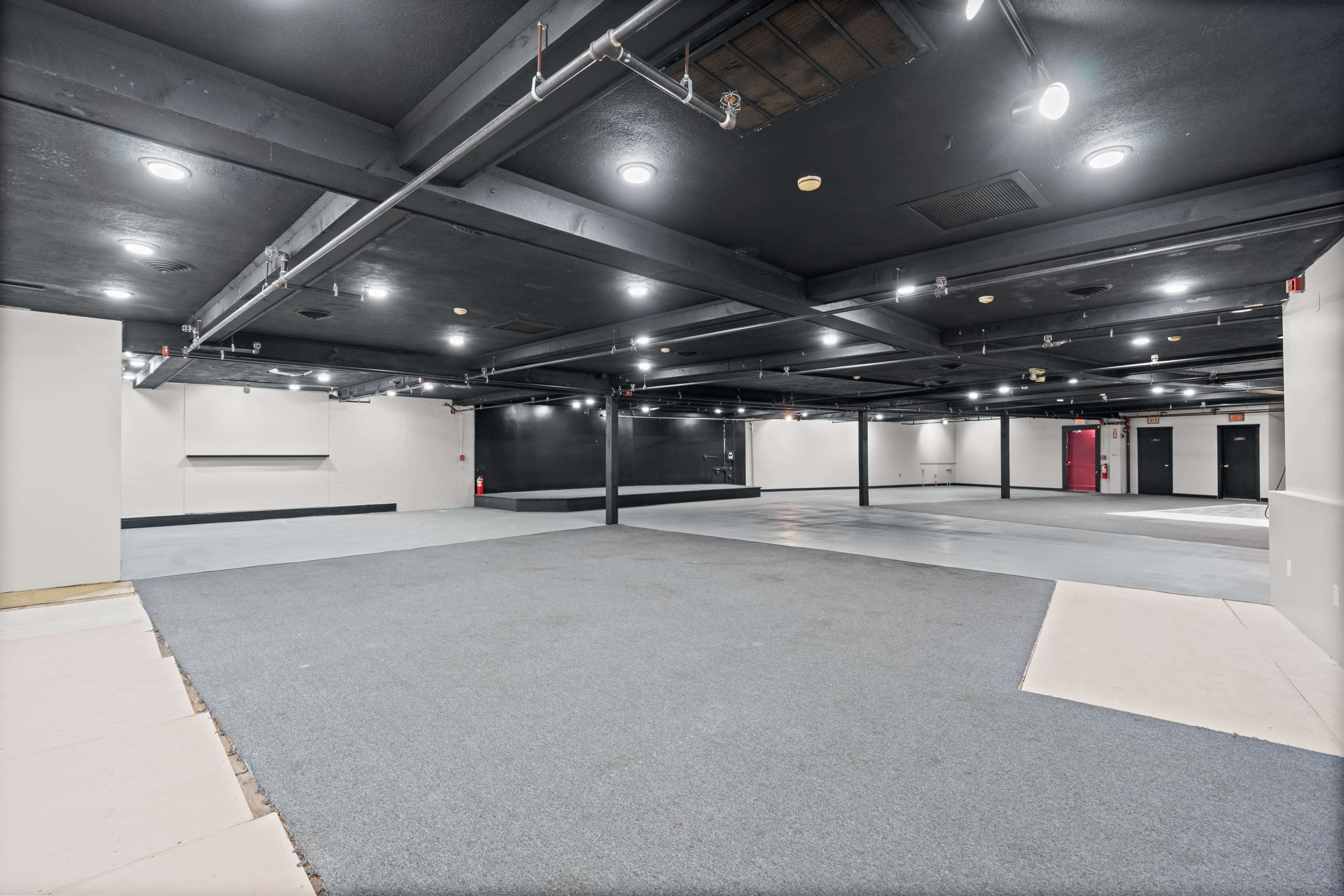 The image shows a spacious, well-lit interior with black ceilings and concrete floors, featuring a mix of carpeted and bare areas.