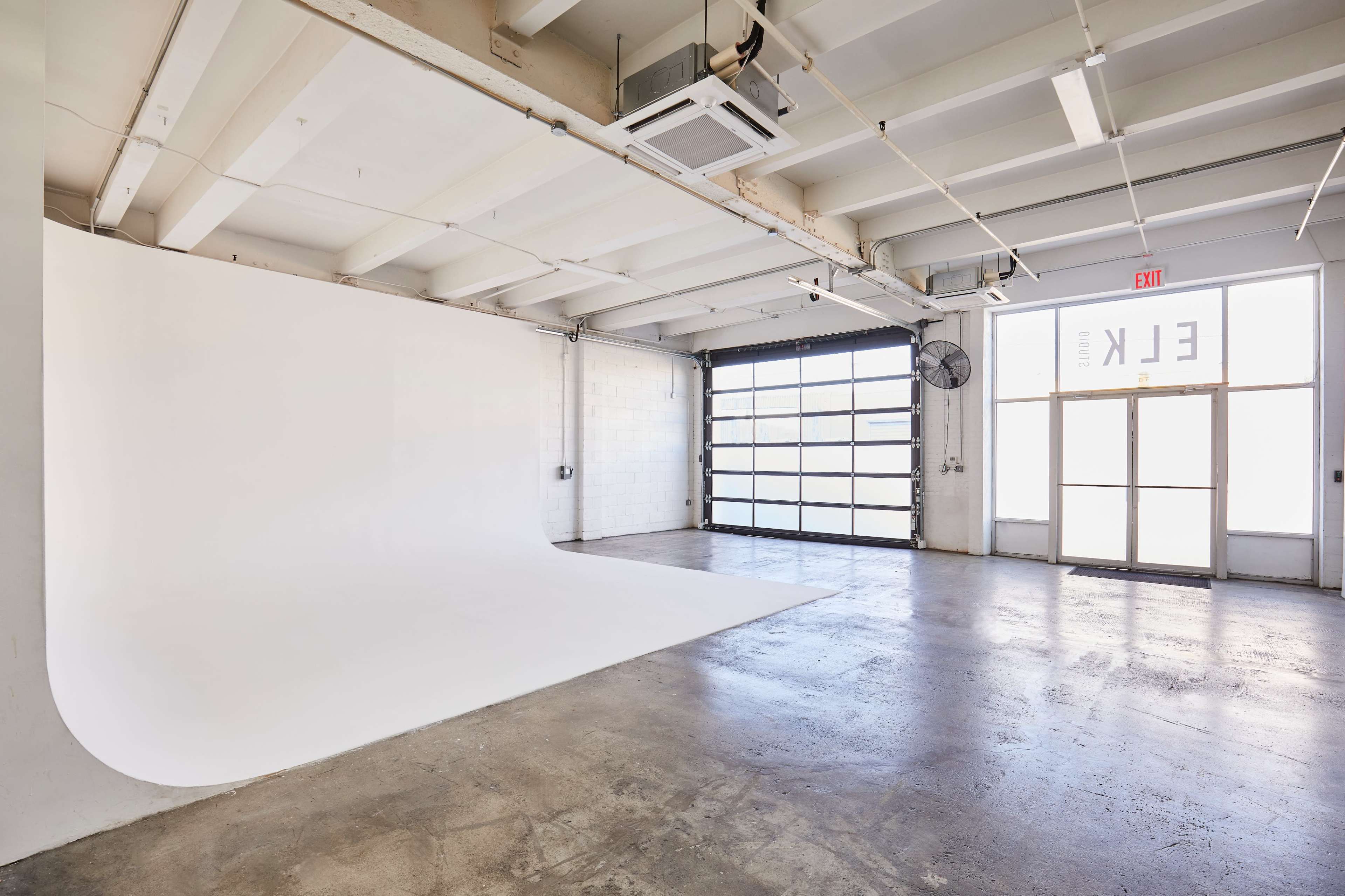 A spacious studio with a white backdrop and large windows, featuring a concrete floor and exposed beams.