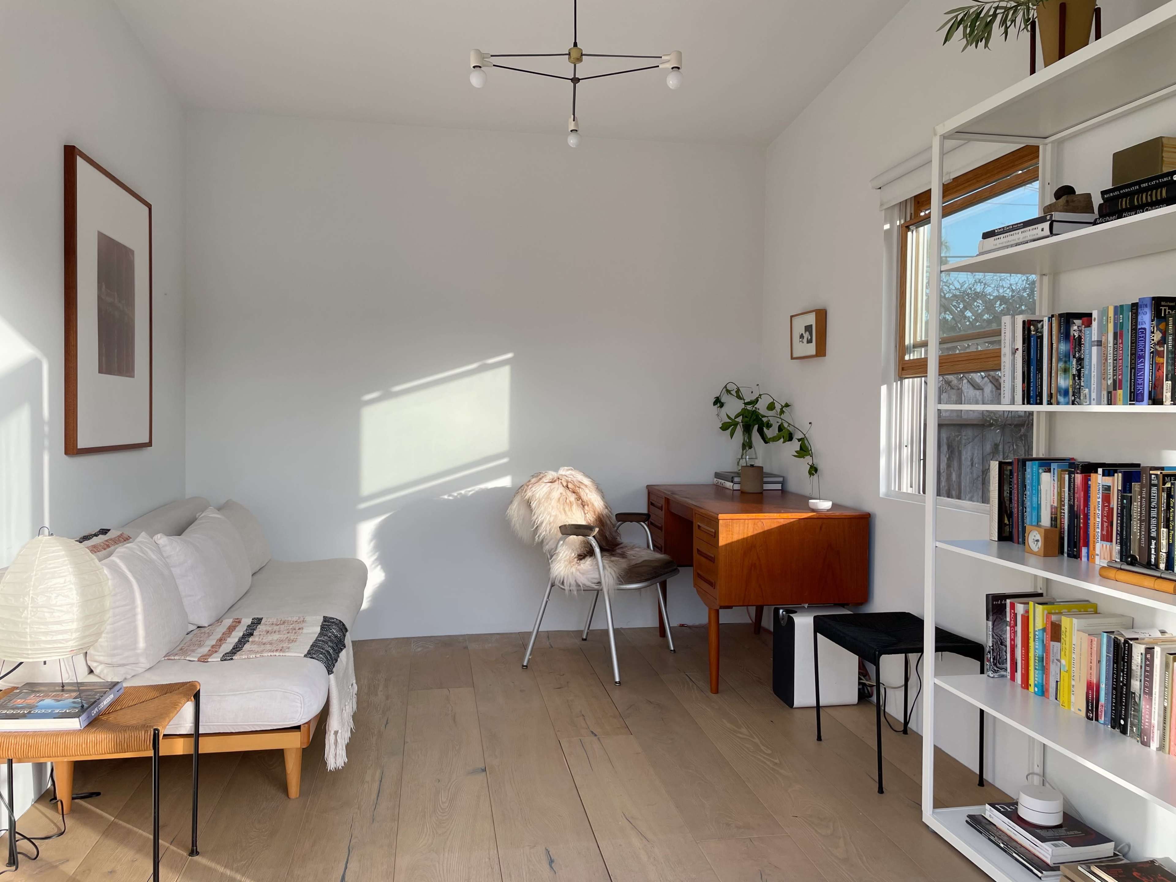 A bright, minimalist living room with a white sofa, a wooden desk, and a bookshelf filled with books.