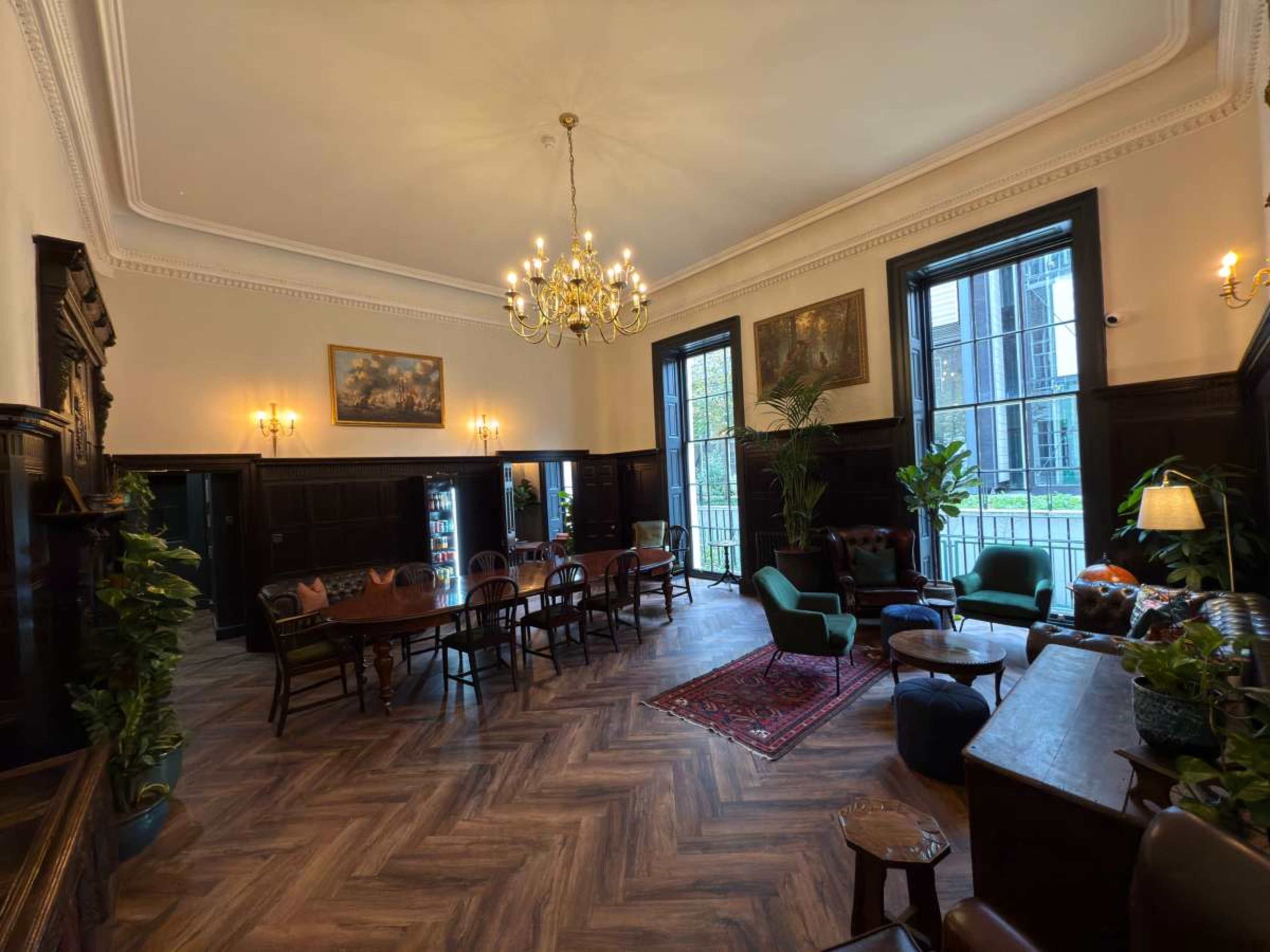The image shows a spacious, elegantly decorated lounge area with a chandelier, wooden furniture, and large windows, surrounded by plants.