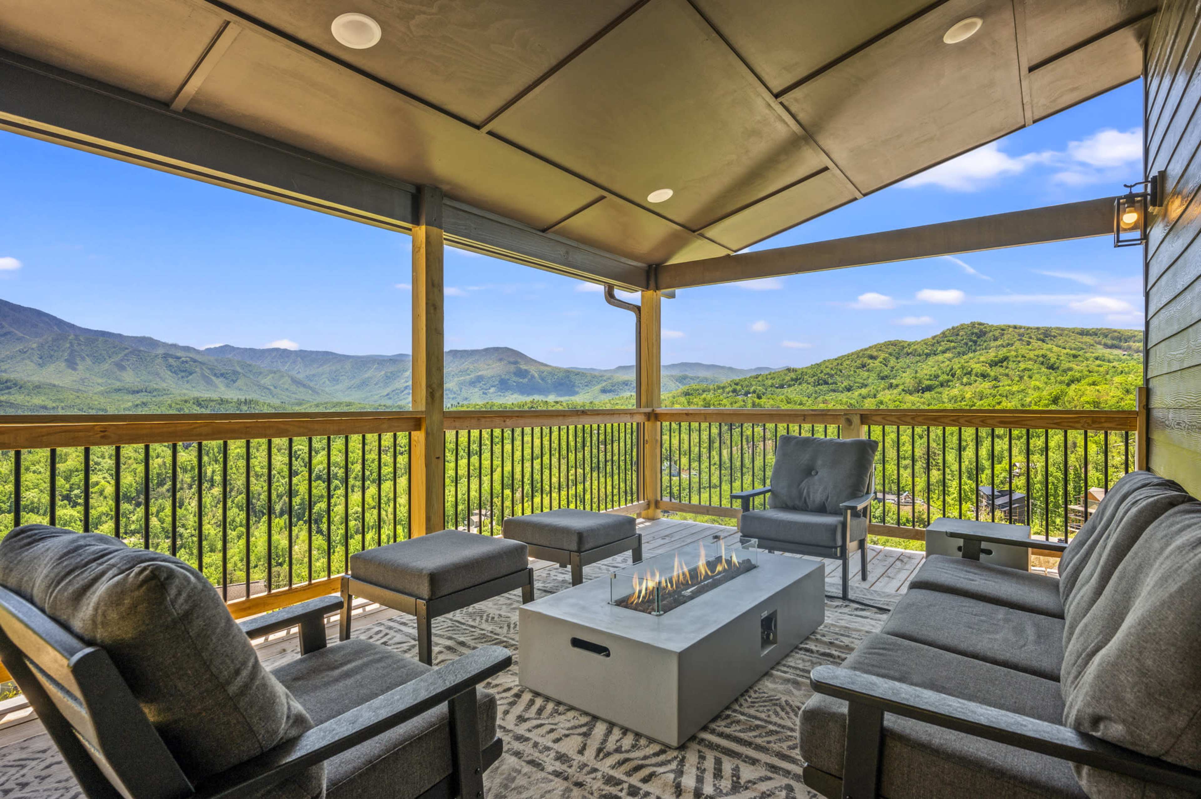 A covered balcony features comfortable seating and a modern fire pit, overlooking a lush mountain landscape.