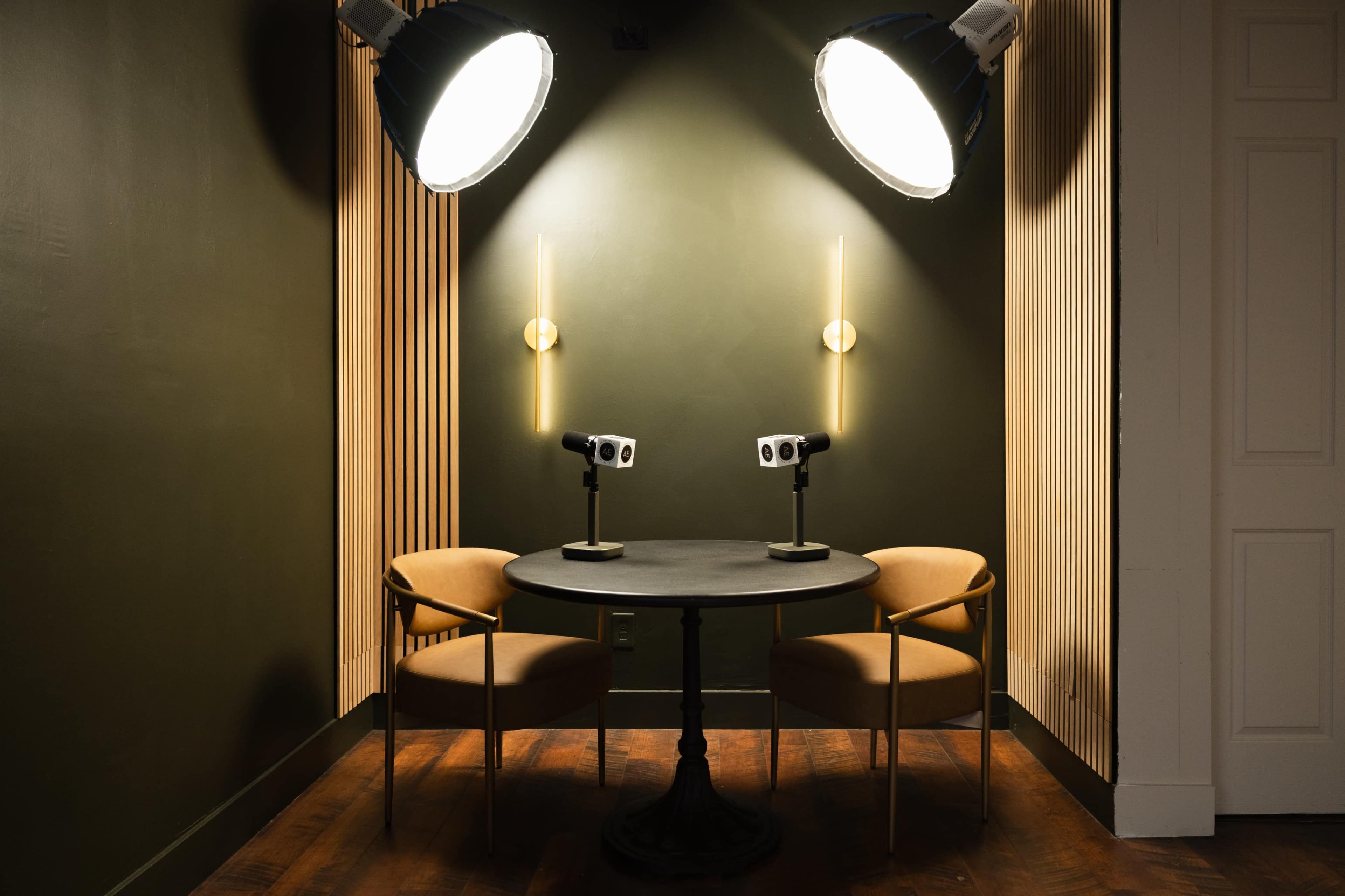 A small, well-lit seating area features two chairs and a round table, with two cameras positioned on stands facing the chairs.