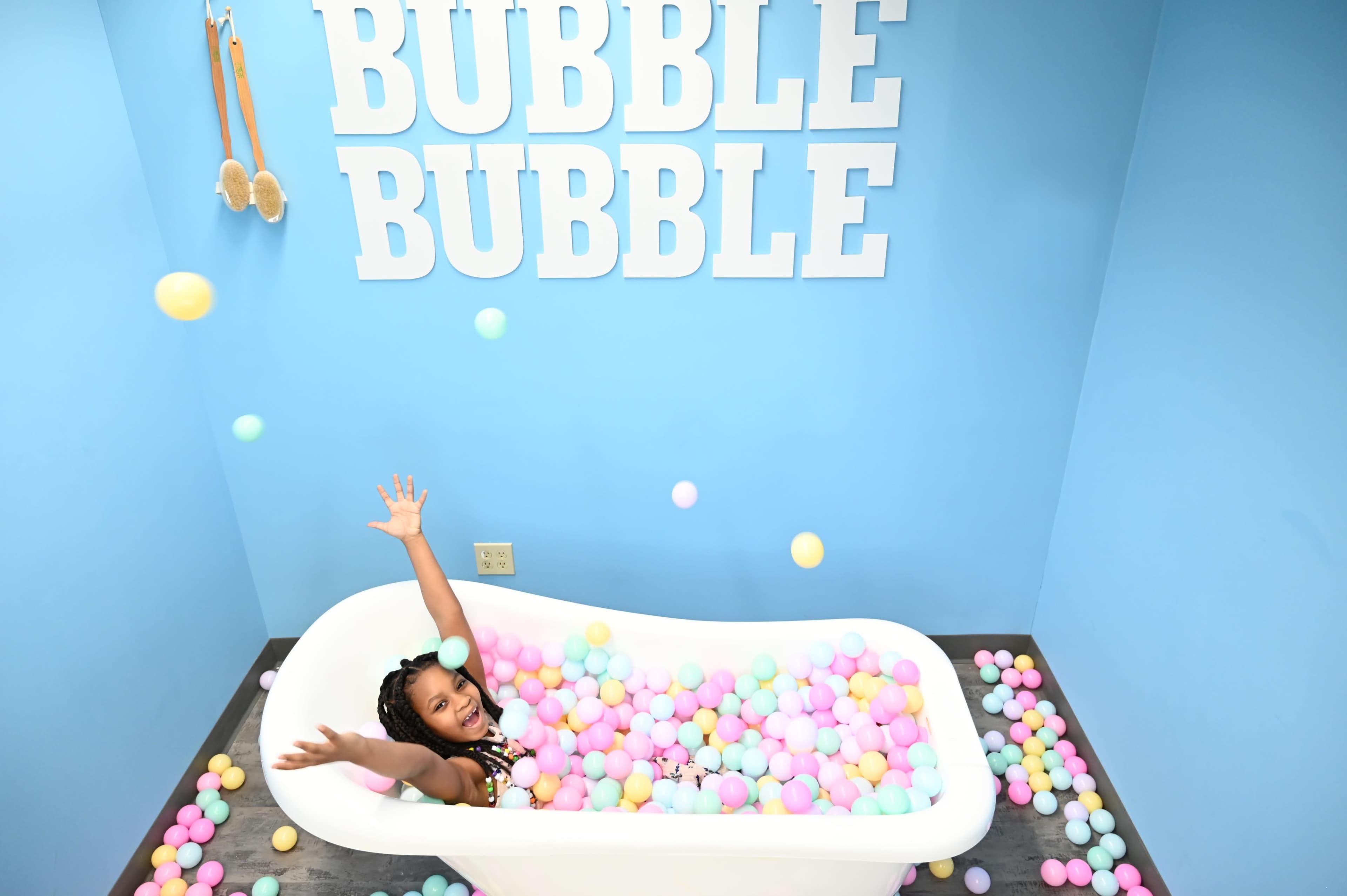 A girl sits in a white bathtub filled with colorful balls, surrounded by blue walls with the words "BUBBLE BUBBLE" displayed above her.