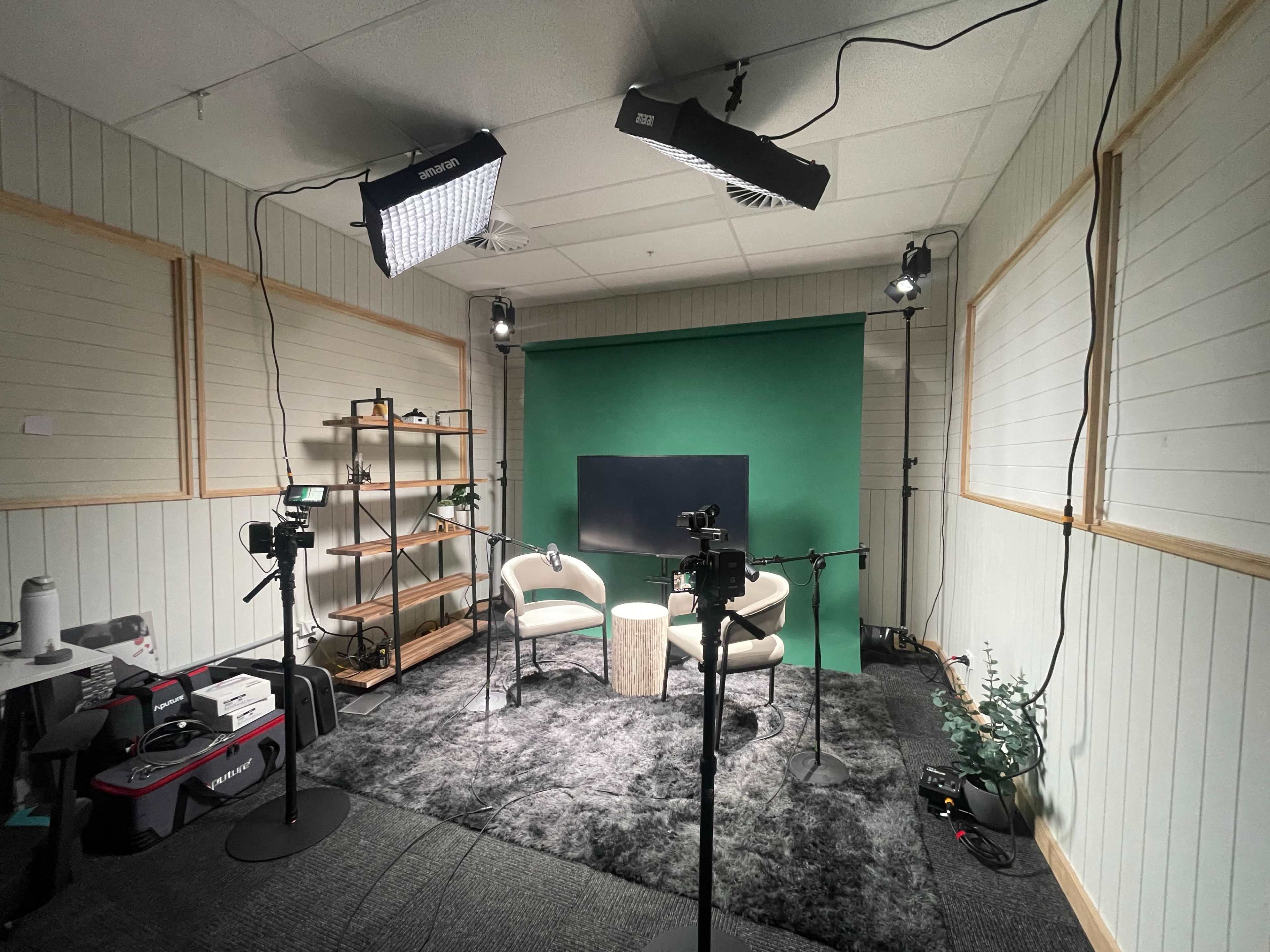 The image shows a video recording studio set up with two chairs, a small table, a green backdrop, and multiple lights positioned around the room.