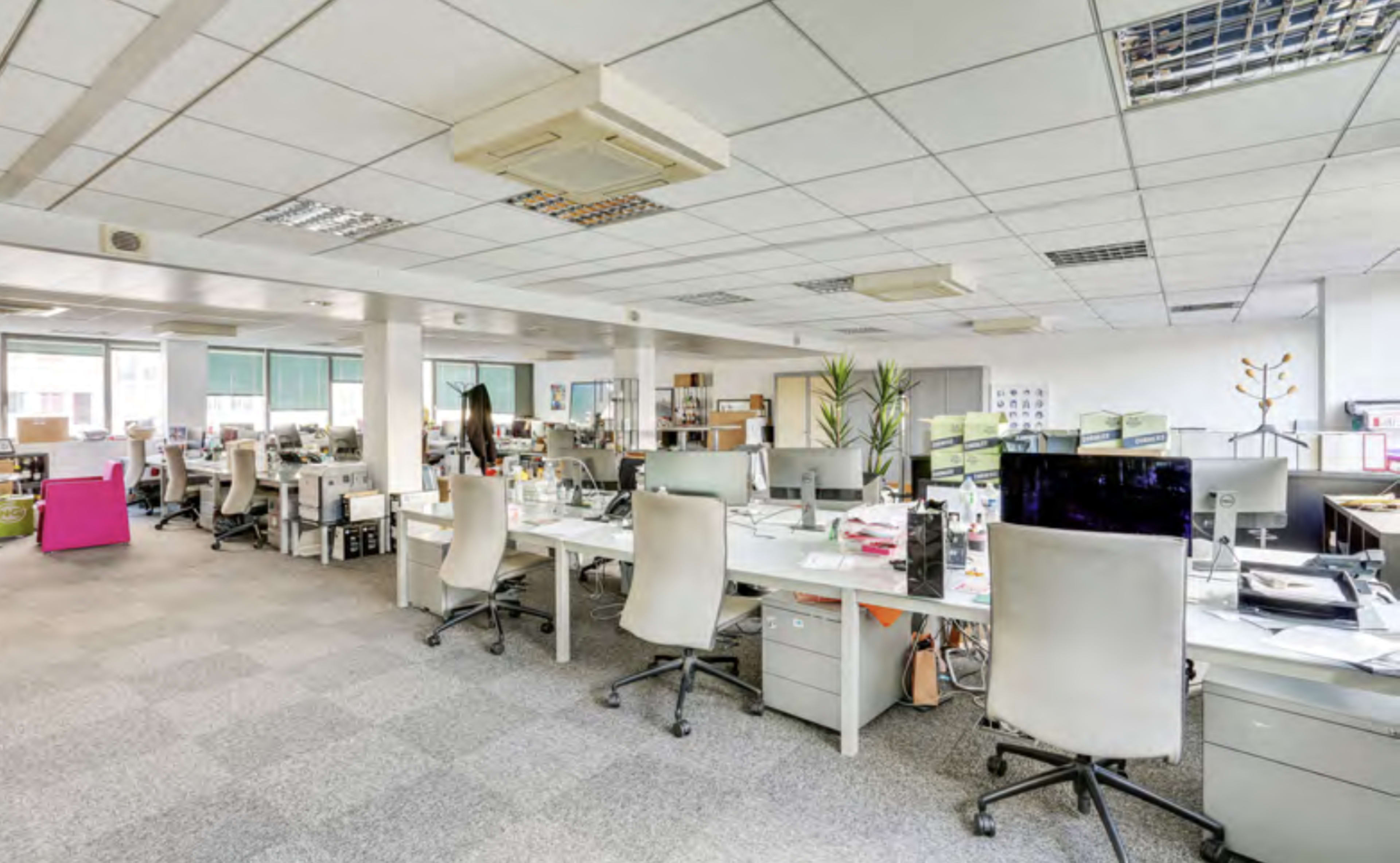 An open office space features several workstations with desks, computers, and chairs arranged in a grid layout.