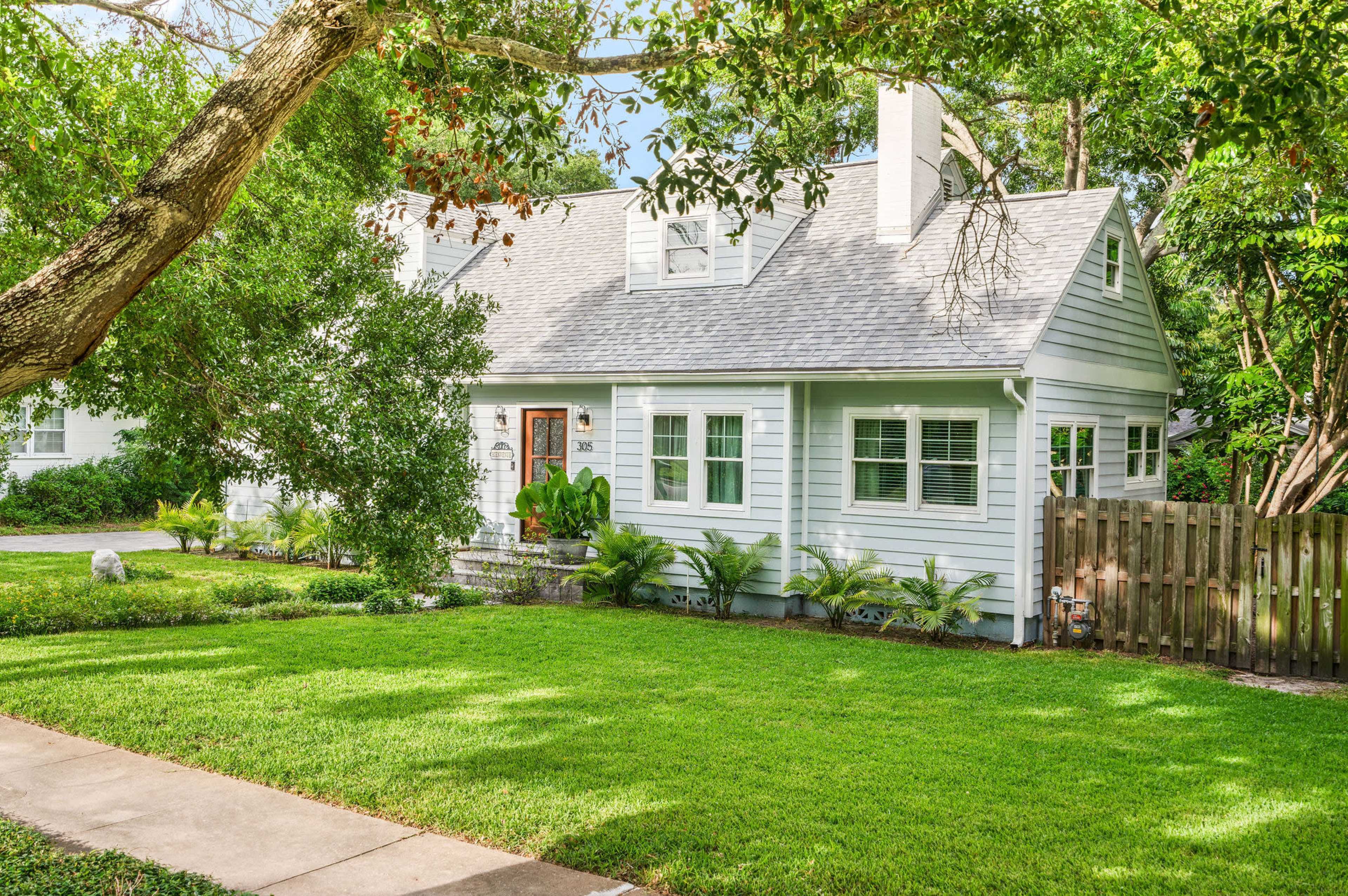 The image shows a light-colored, two-story house surrounded by lush green grass and trees, with a wooden fence and a pathway leading to the entrance.
