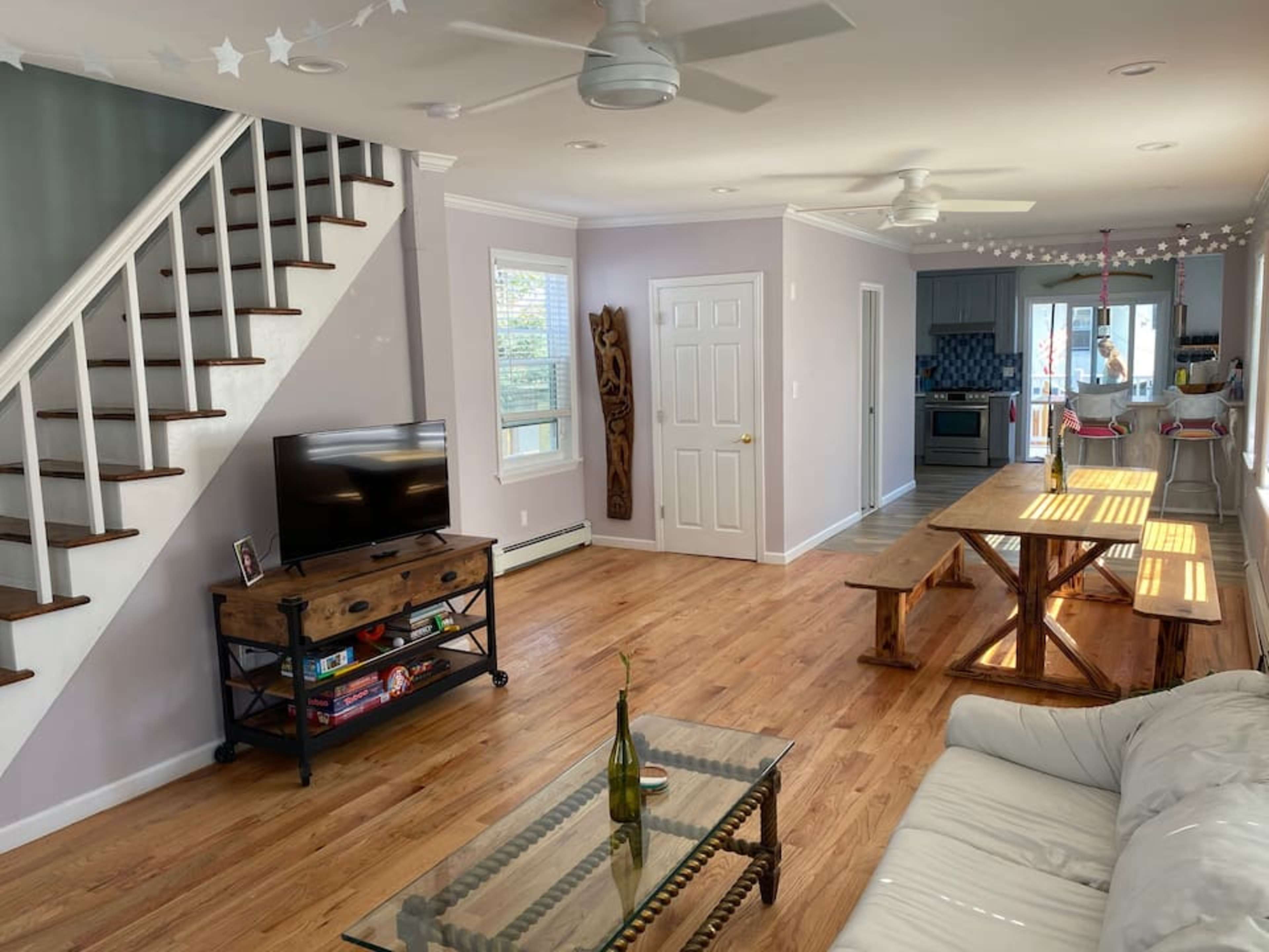 A spacious living area features a wooden dining table, a sectional sofa, a television on a stand, and stairs leading to the upper floor.