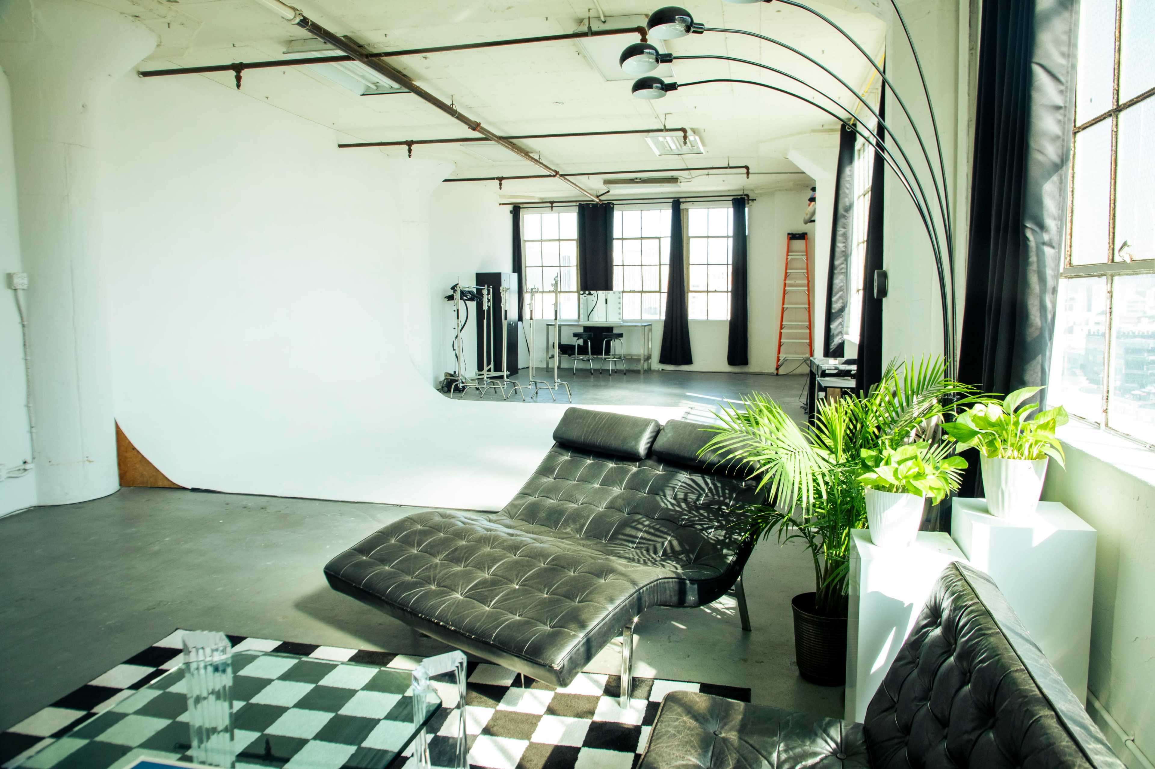 The image shows a spacious, contemporary studio with a black lounge chair, a checkerboard coffee table, and a large window providing natural light.