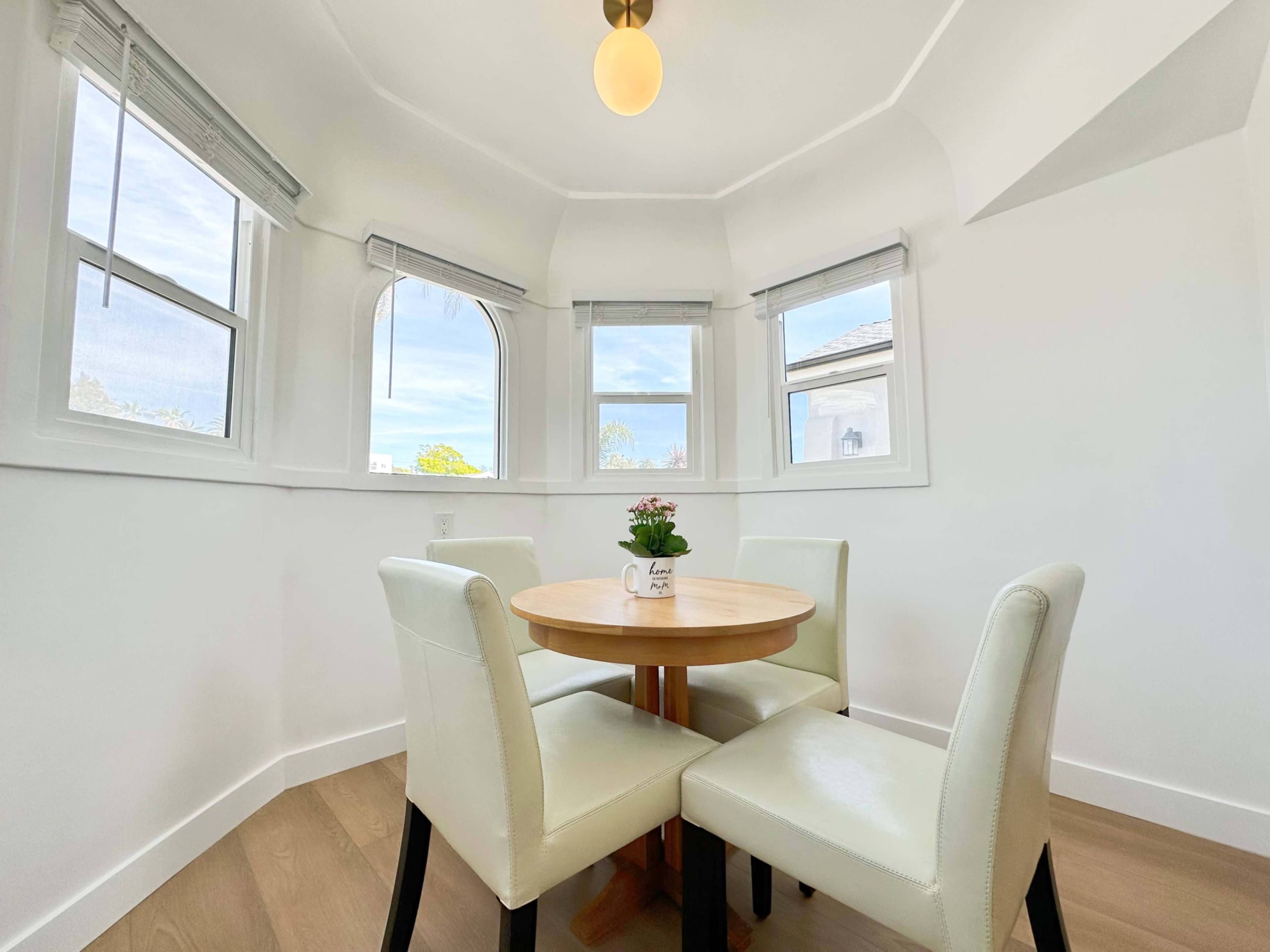 A small round wooden table with two white leather chairs is placed in a bright, windowed corner of a room.