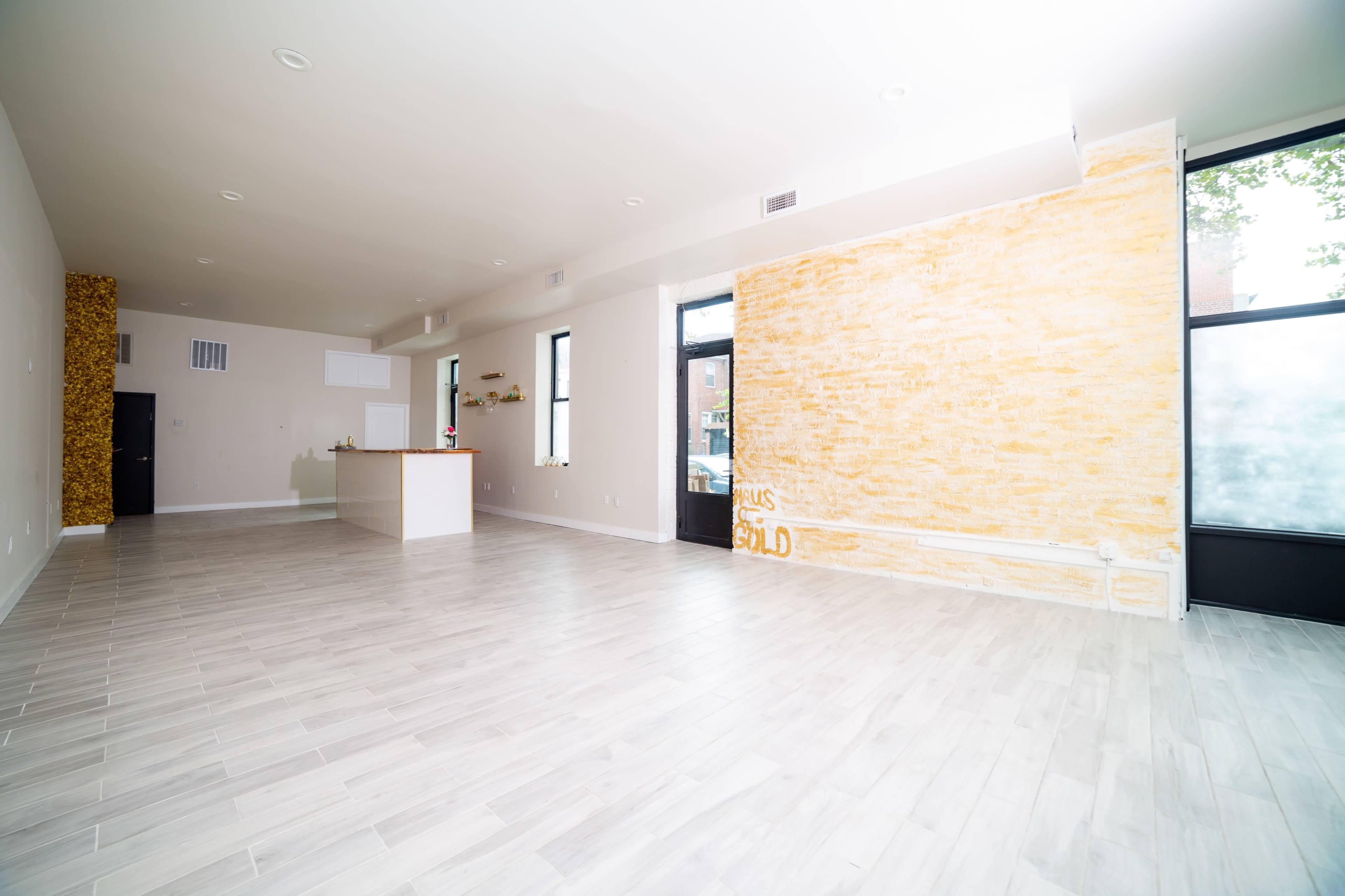 The image shows an interior space with light-colored flooring, a yellow feature wall, a kitchen area, and large windows.