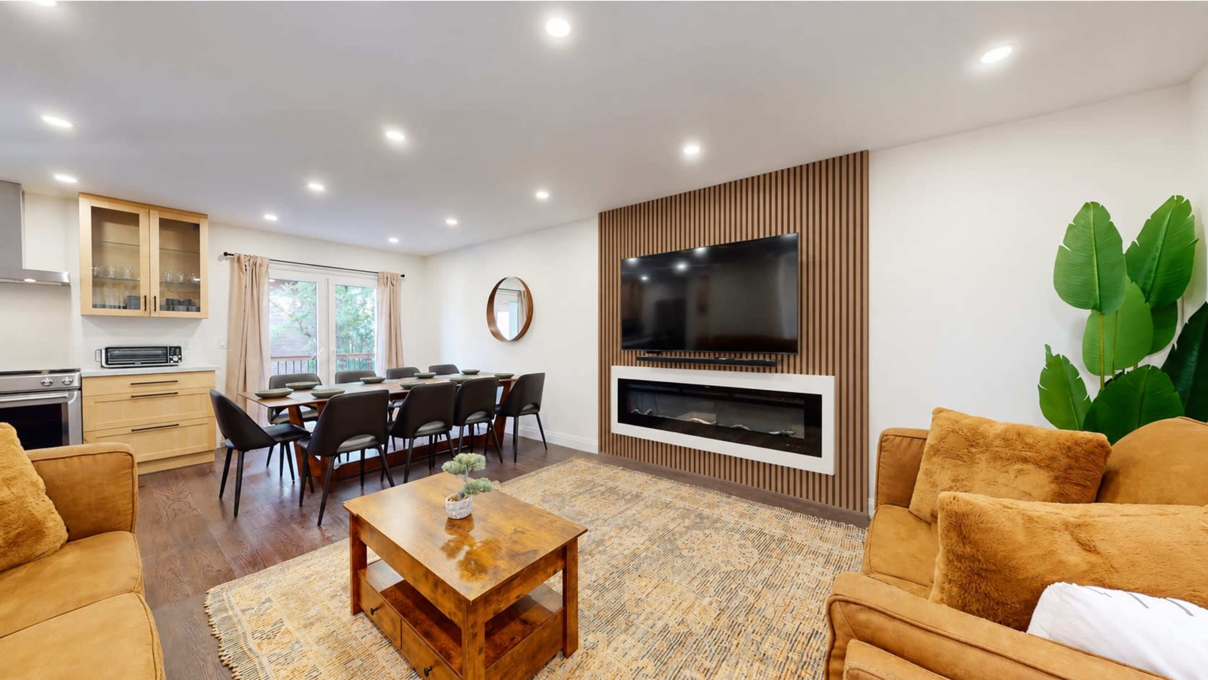 A modern living room features a sofa and armchairs, a wooden coffee table, a dining area with a table and chairs, and a wall-mounted television above a fireplace.