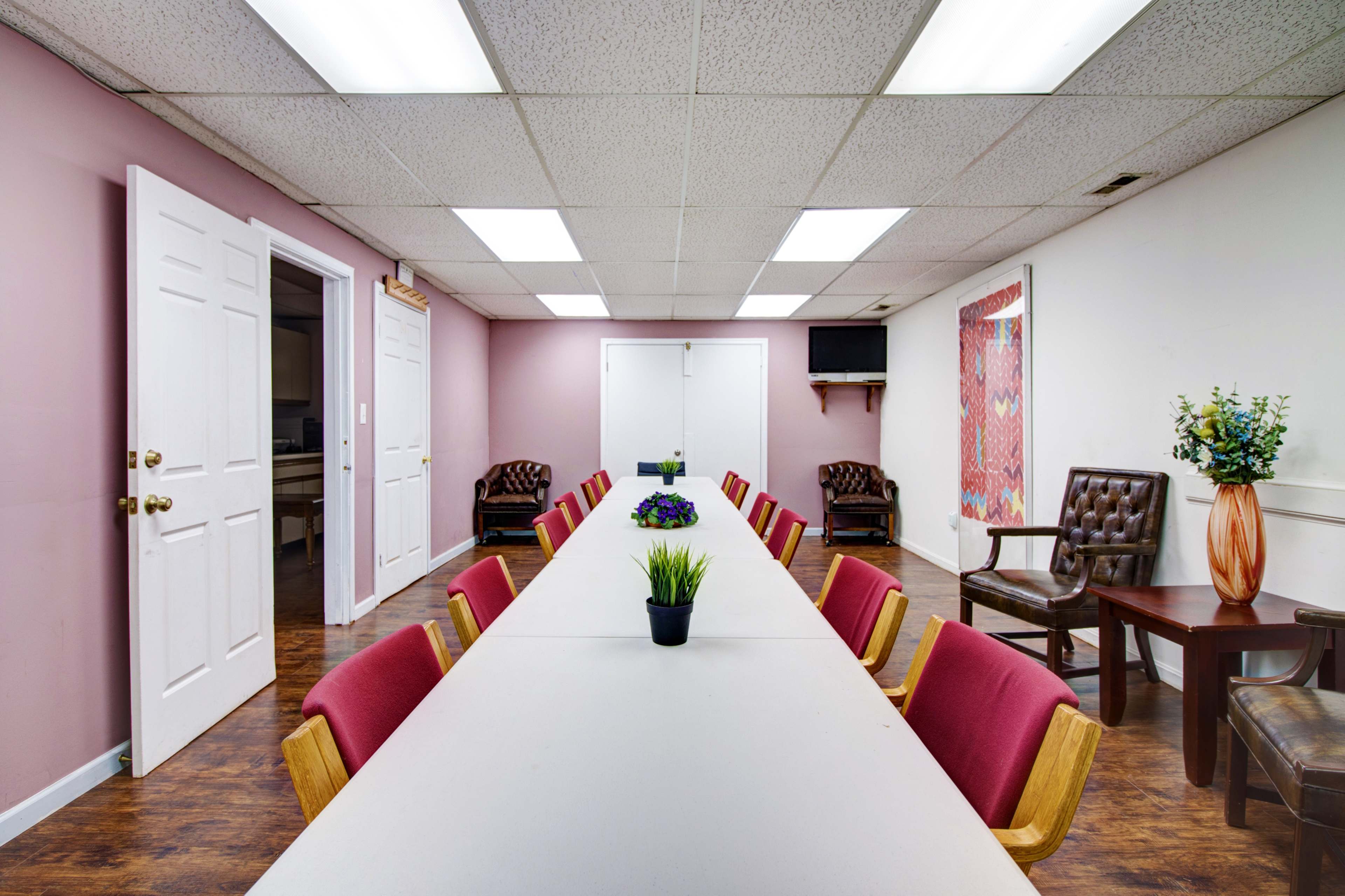 A meeting room with a long table surrounded by chairs, flanked by two doors and a wall-mounted TV.