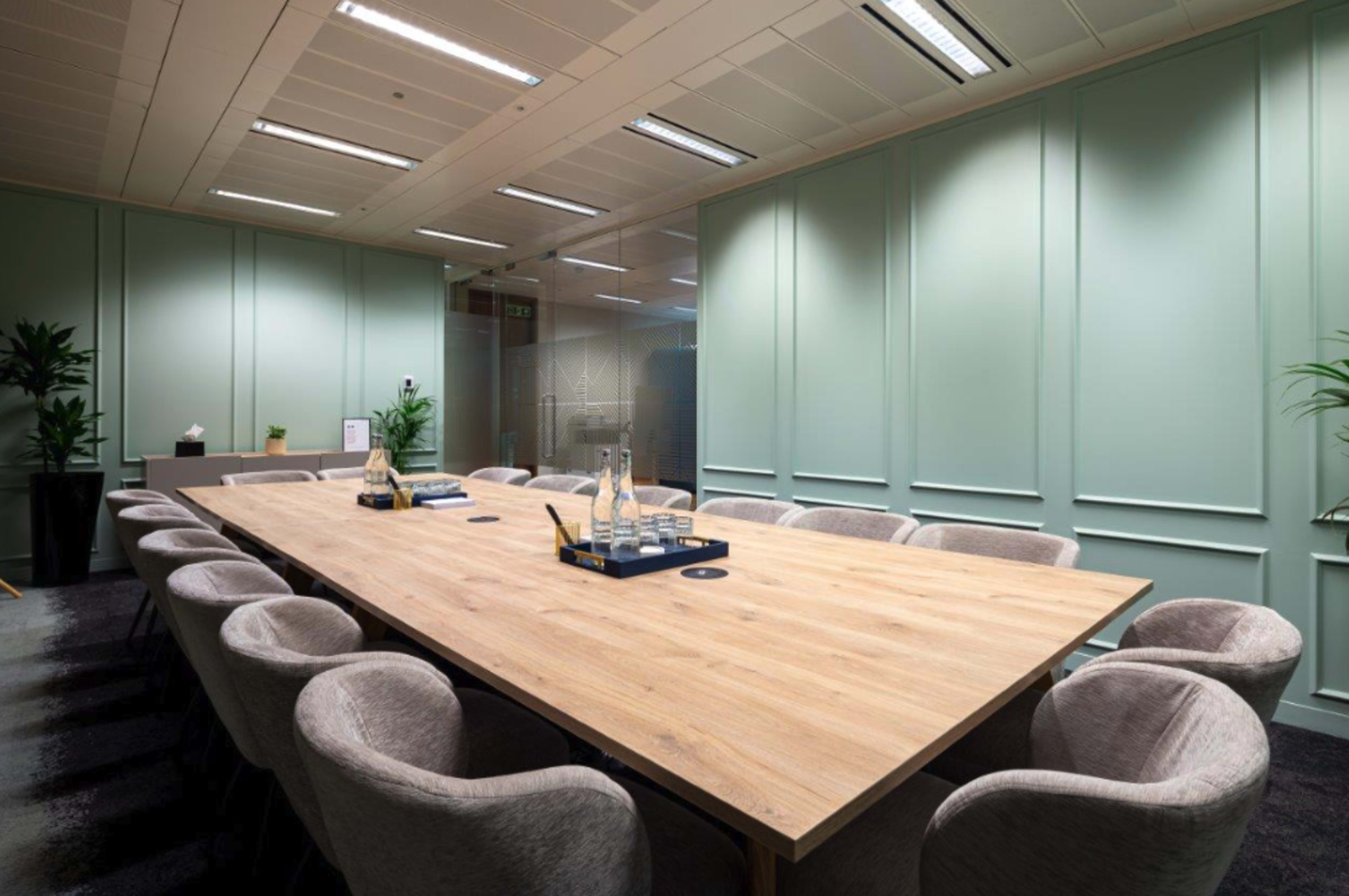 A long wooden conference table is surrounded by cushioned chairs in a well-lit meeting room with green walls and potted plants.