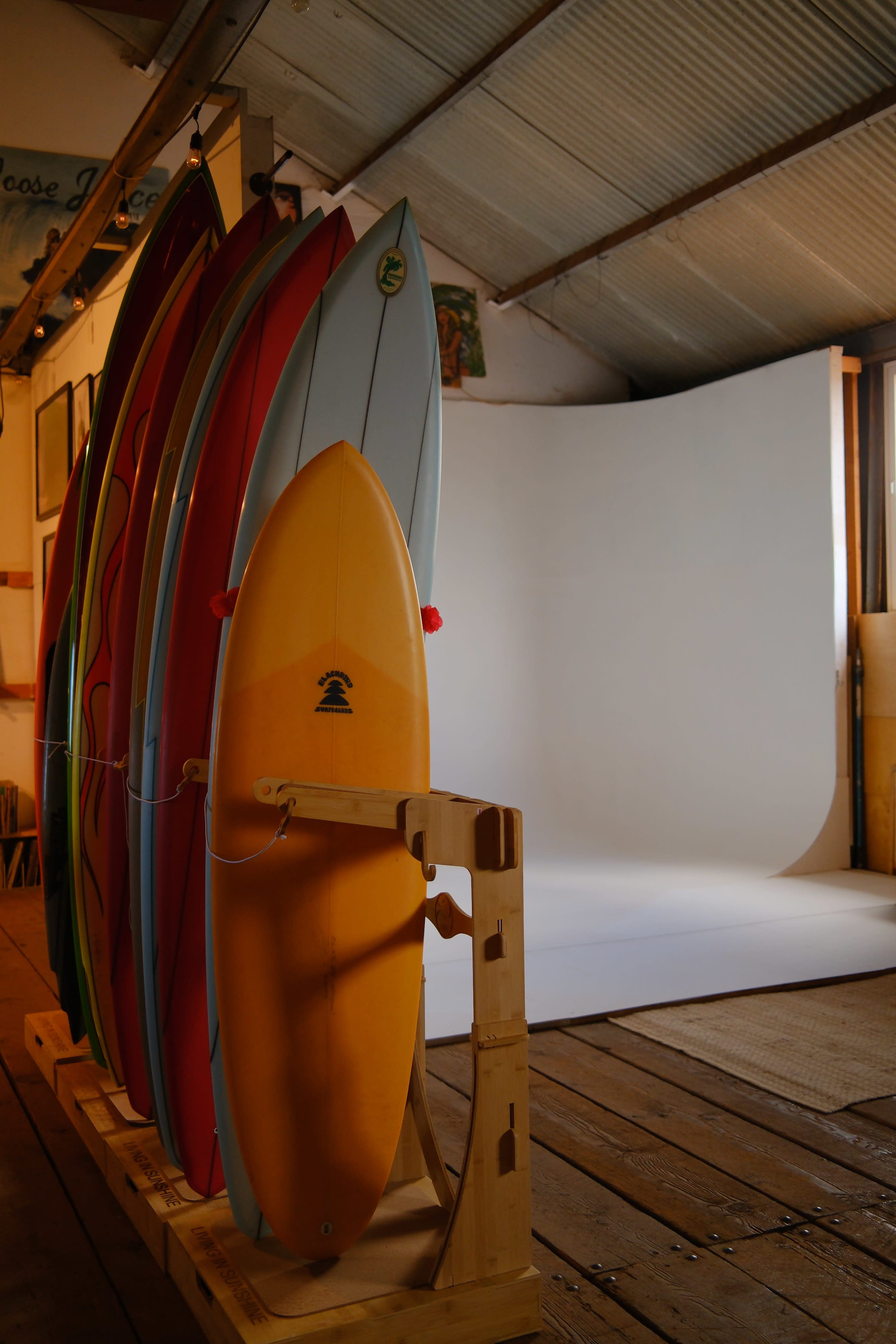 Six surfboards are leaning against a wooden rack beside a blank white backdrop in a room with wooden flooring.