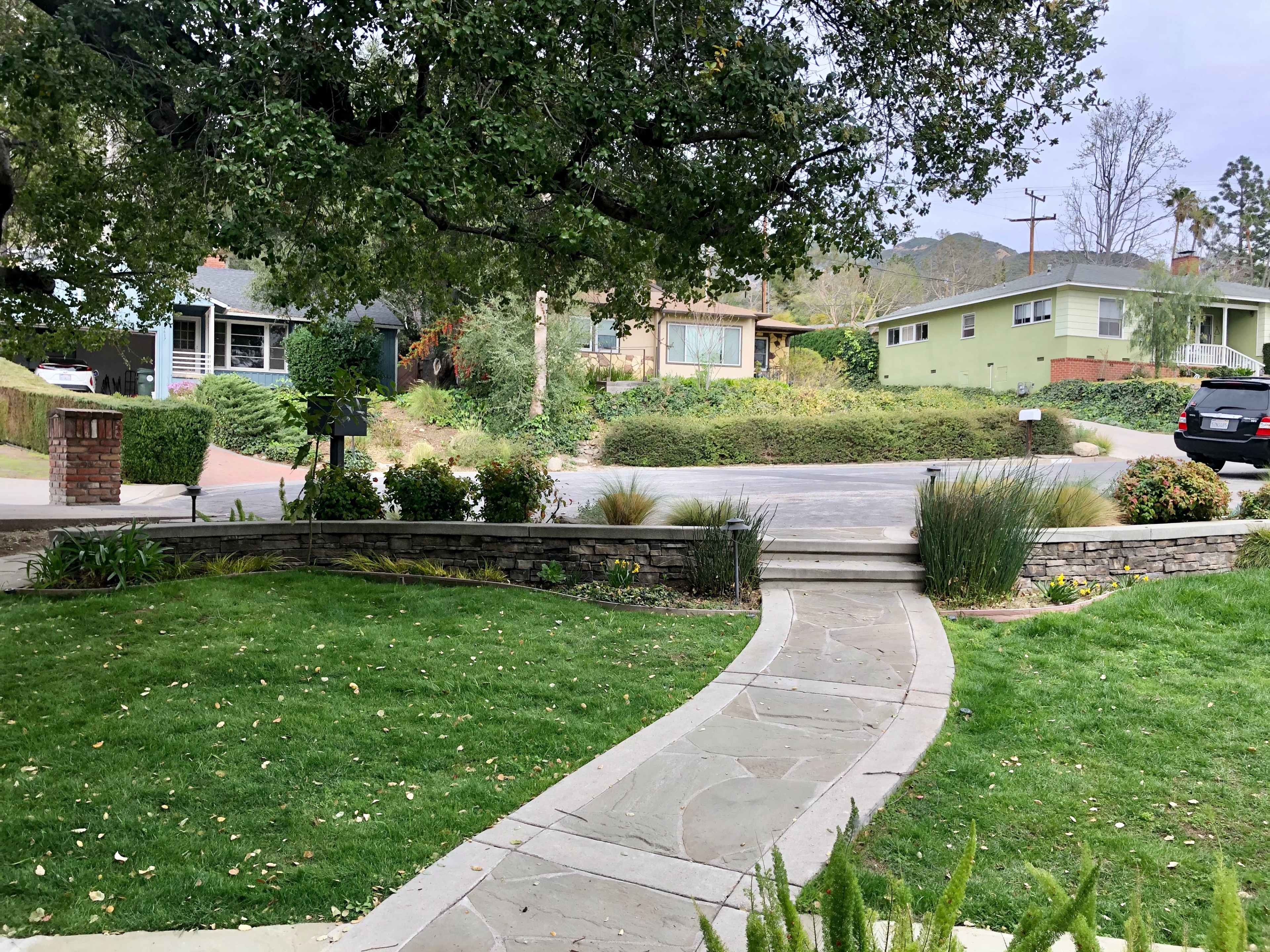 The image shows a landscaped front yard with a curved stone pathway leading to a road, bordered by houses and greenery.