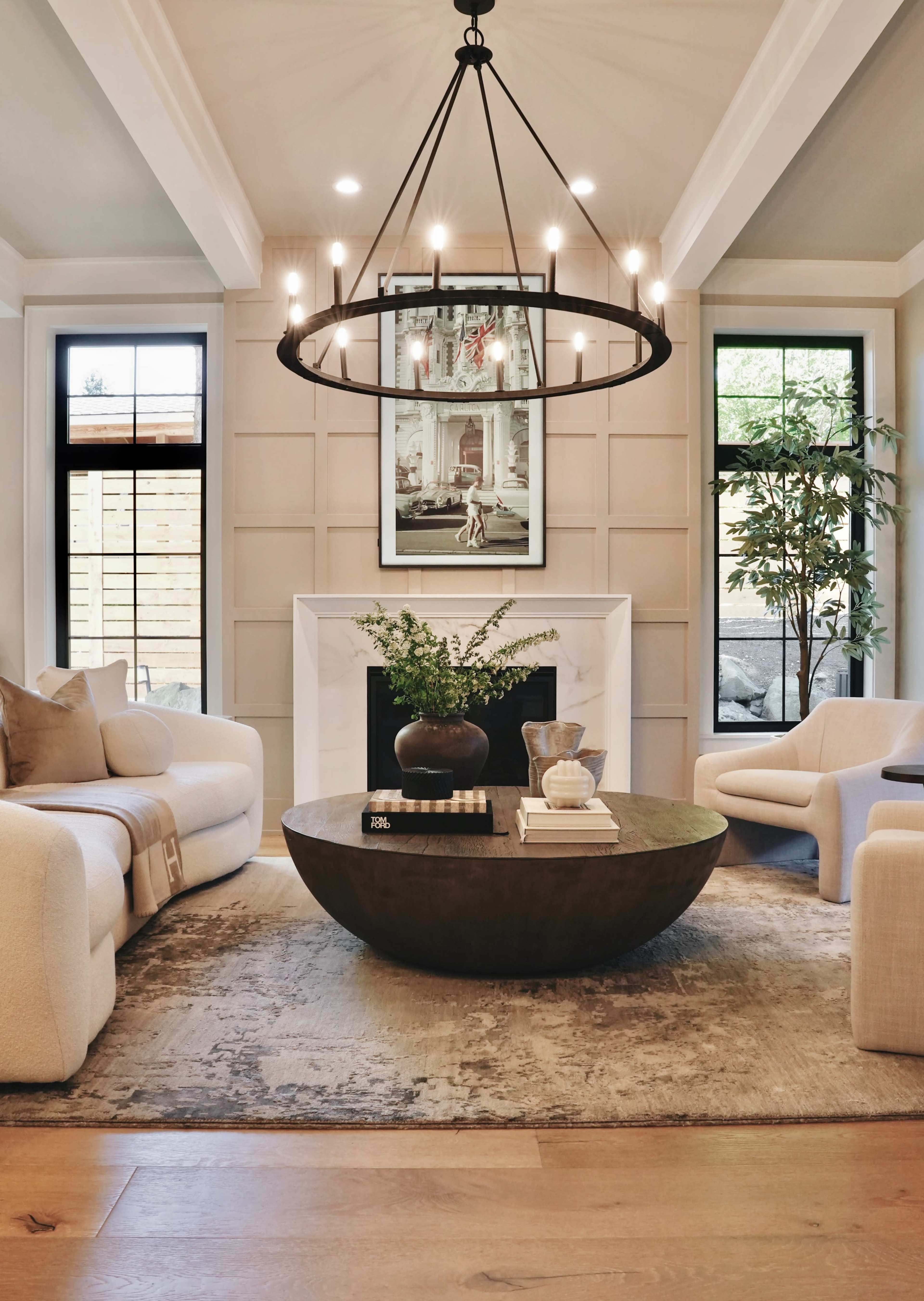 The image shows a bright living room featuring two upholstered chairs, a round coffee table, a large chandelier, and artwork above a fireplace, with windows that provide natural light.