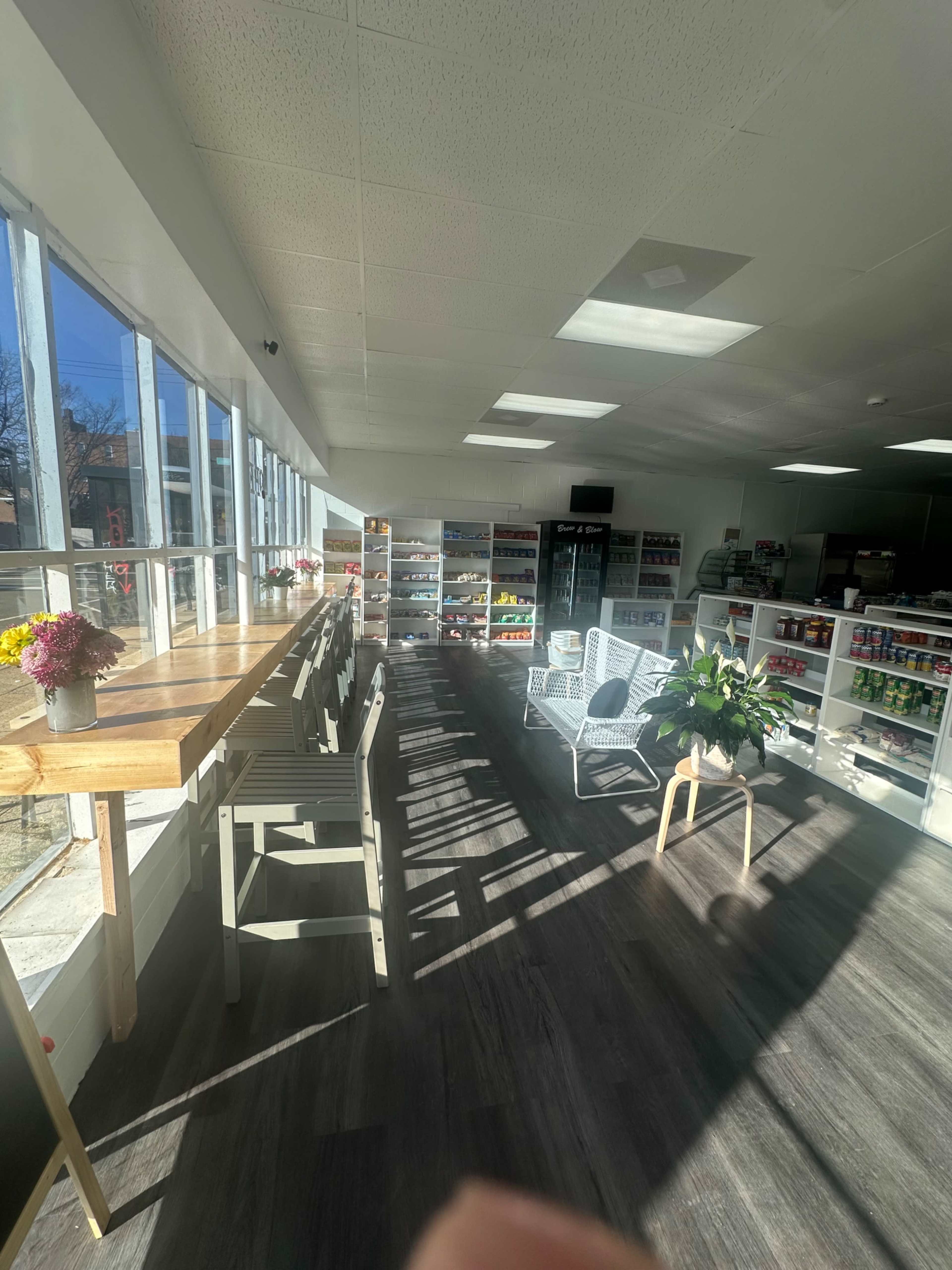 The image shows a bright, spacious interior with a long wooden table, white chairs, and shelves filled with various products.