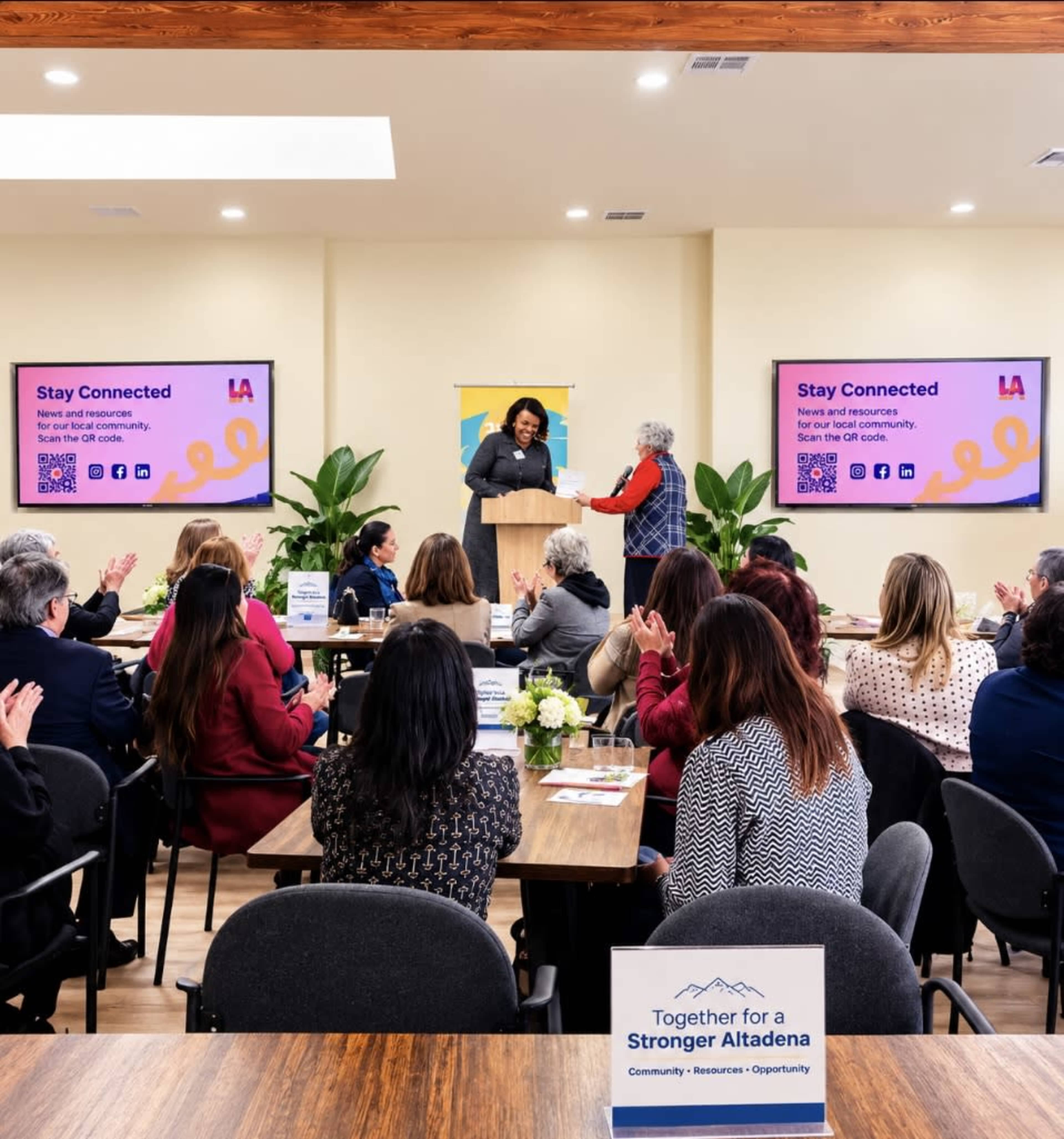 A group of people are seated around tables in a conference room, watching a presentation while some attendees applaud a speaker receiving an award.