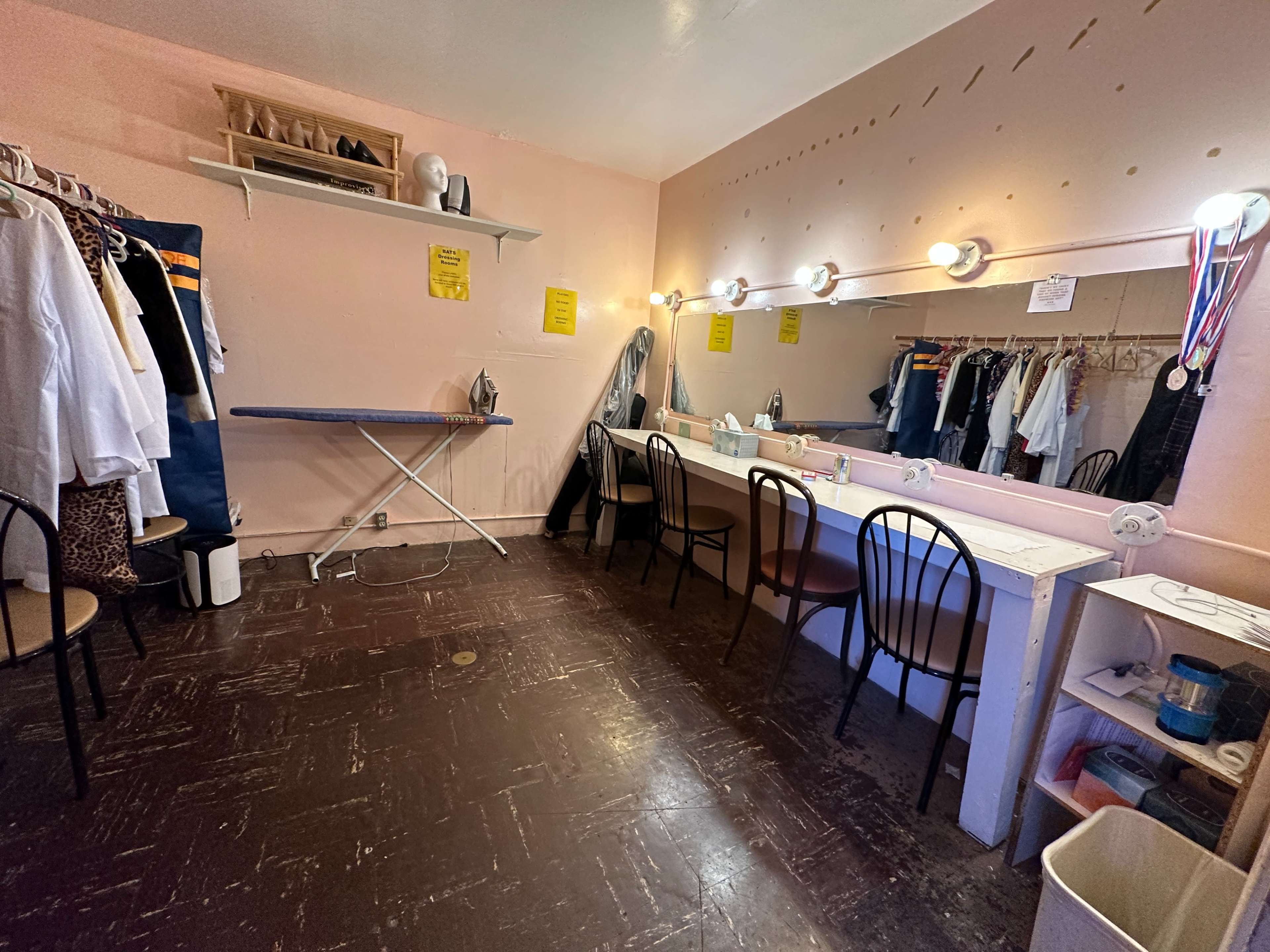 The image shows a dressing room with a row of mirrors, lights, and several chairs, as well as clothing hanging on a rack and an ironing board.