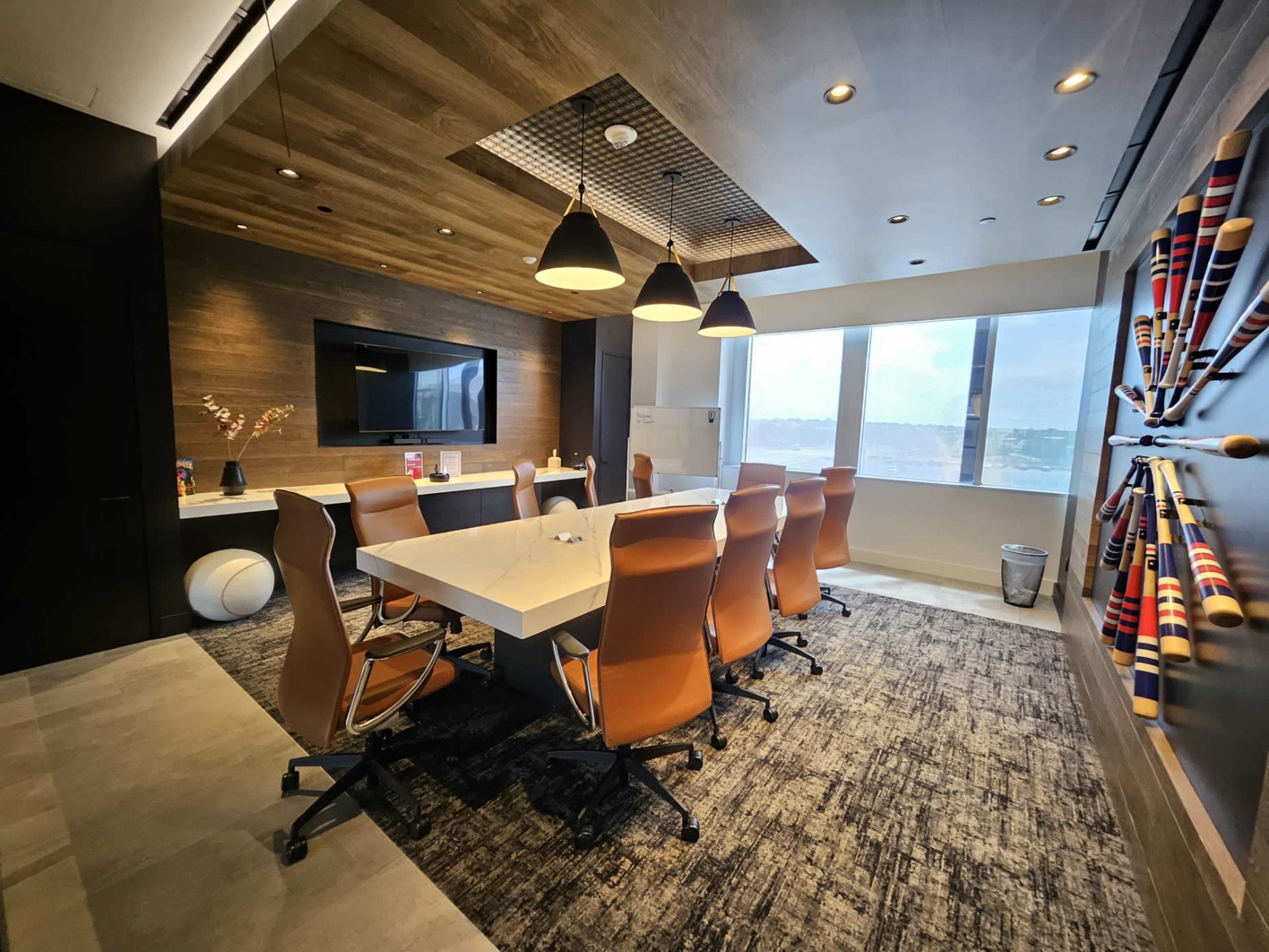 A modern conference room features a large table surrounded by brown leather chairs, with a television on the wall and decorative baseball bats arranged artfully.