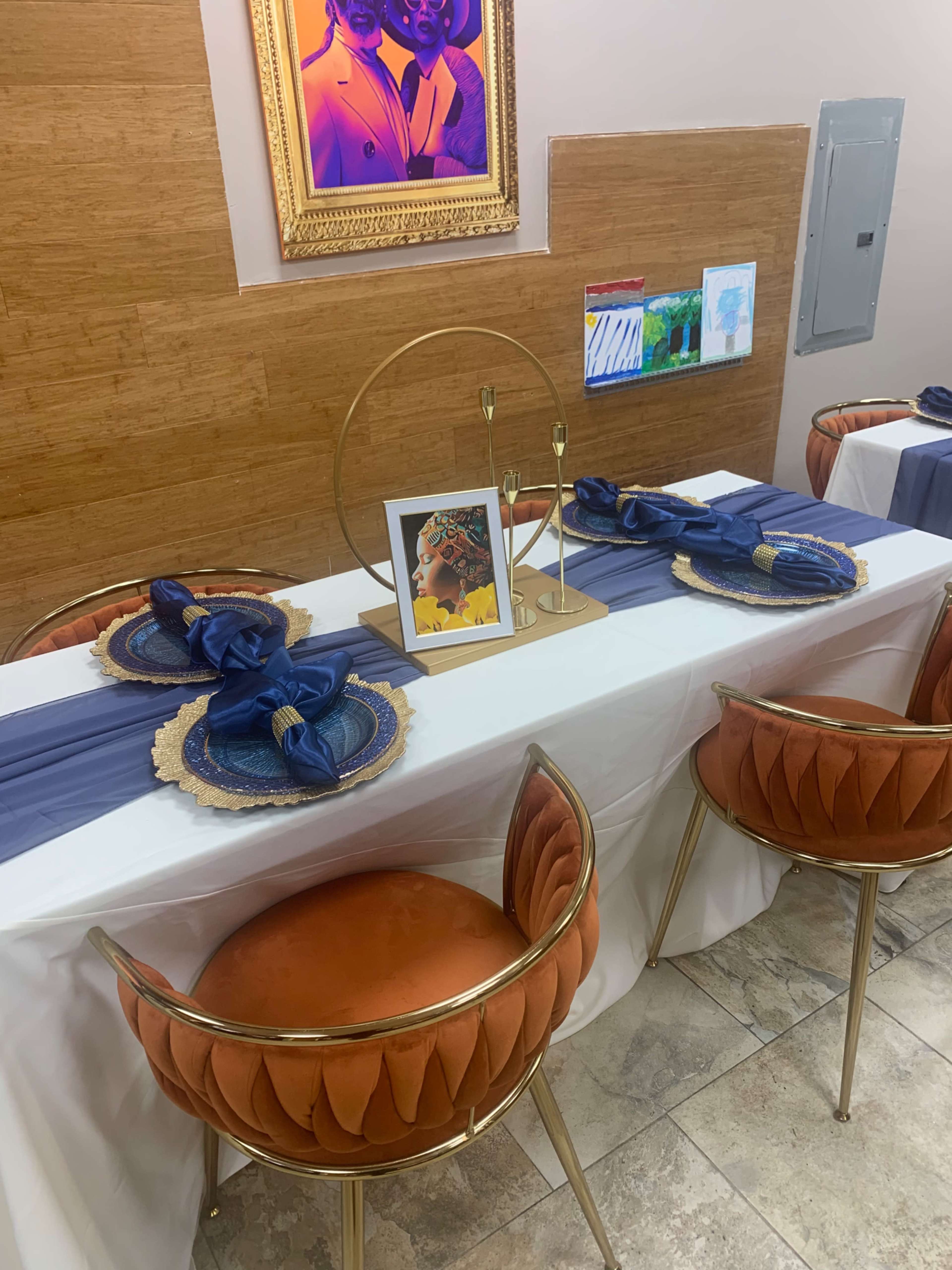 A dining area with two tables elegantly set with navy blue table runners, gold-accented plates, and styled chairs against a wooden backdrop.