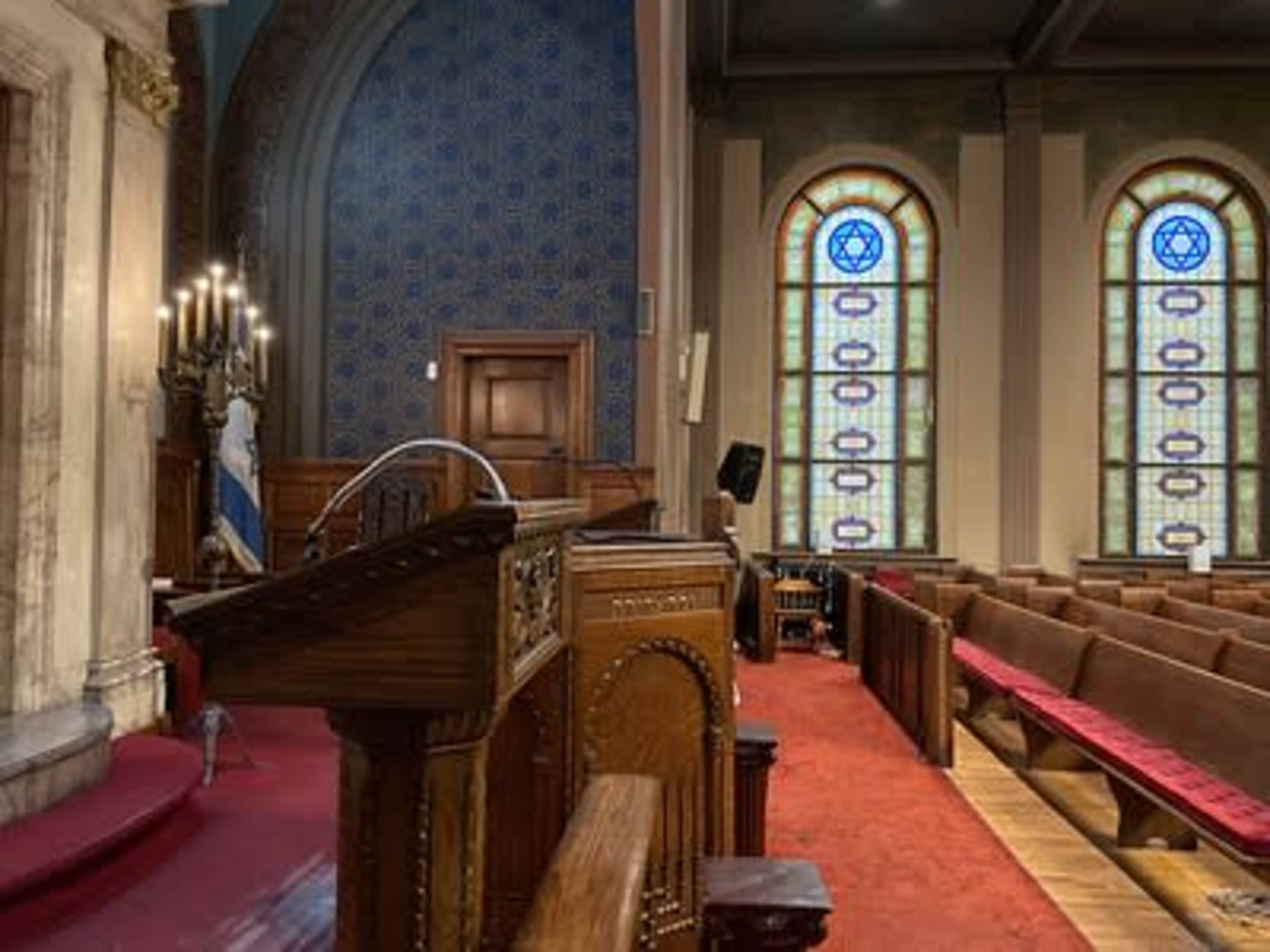 The interior of a synagogue, featuring a wooden podium, rows of pews, and stained glass windows with Star of David designs.