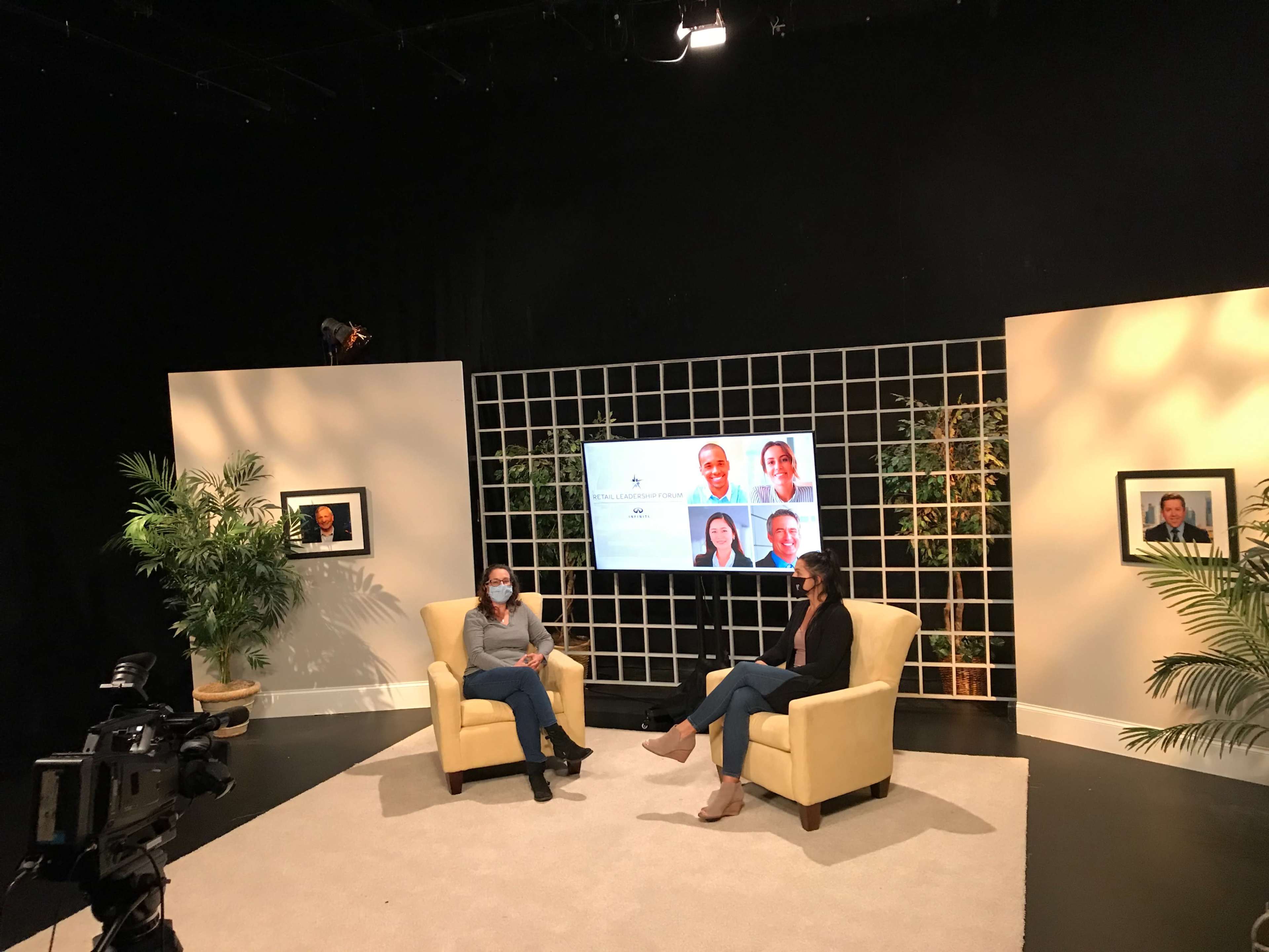 Two women are seated in a television studio set, facing each other, with a large screen displaying headshots in the background and cameras positioned to capture the scene.
