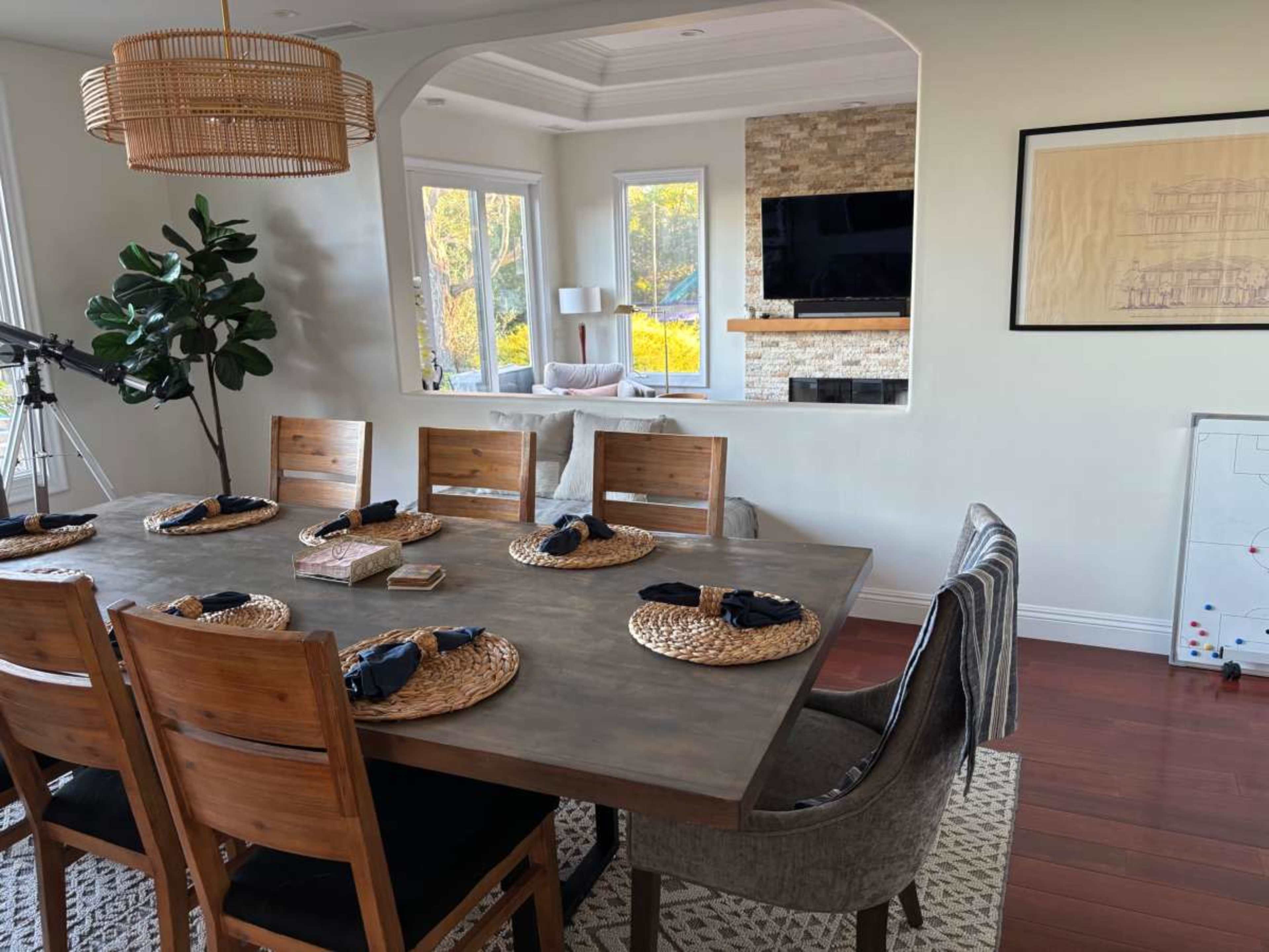 The image shows a dining area with a wooden table set for meals, surrounded by eight chairs, and a cozy living space featuring a television and windows in the background.