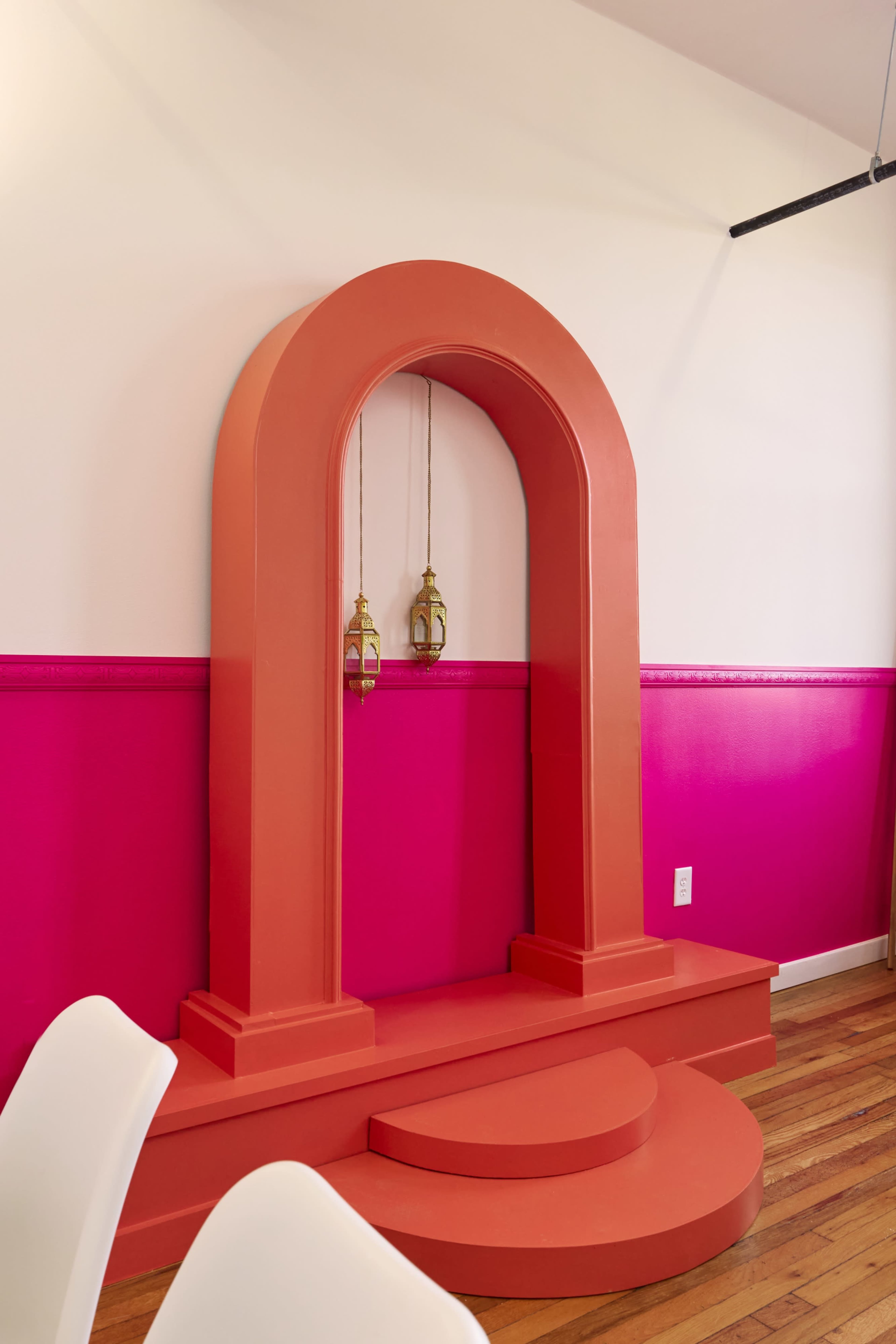 The image shows a stylized architectural feature with an orange arch and a small platform, complemented by two hanging lanterns, set against a backdrop of bright pink walls and wooden flooring.
