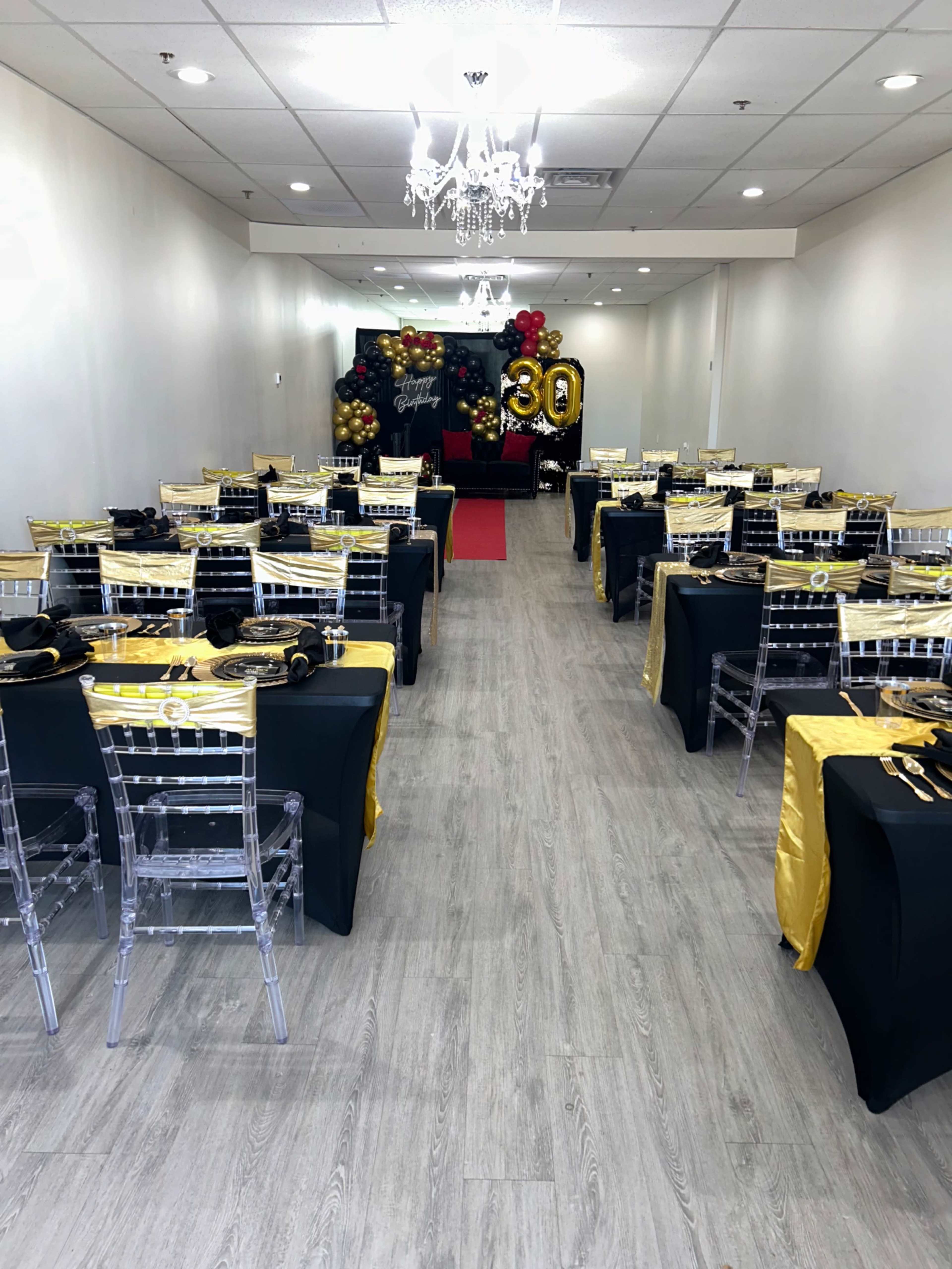 A banquet hall is decorated for a 30th birthday celebration, featuring tables set with black and gold tablecloths, clear chairs, and a red carpet leading to a balloon display.