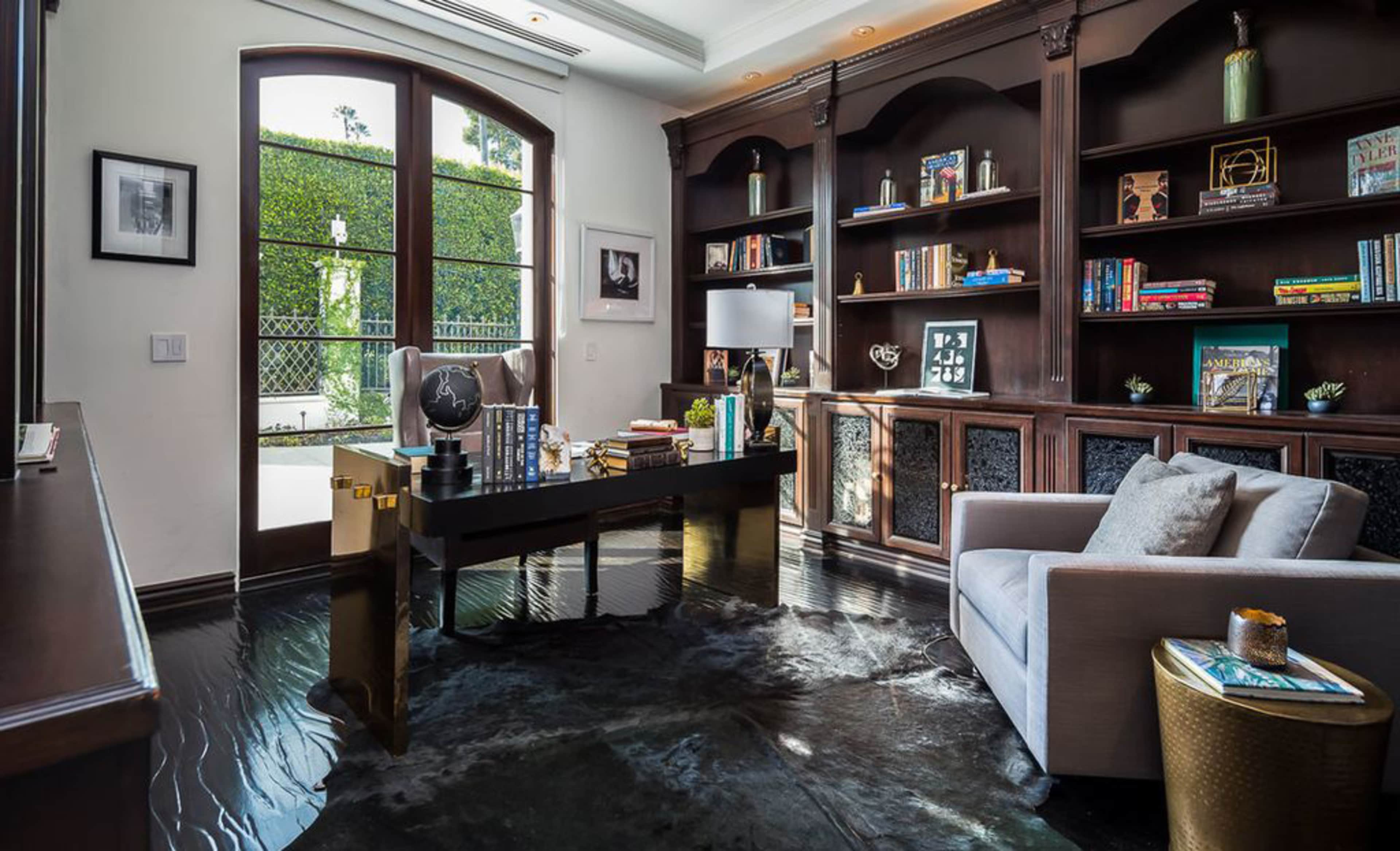 A well-furnished office featuring a wooden desk, bookshelves filled with books, and large glass doors leading to a garden.