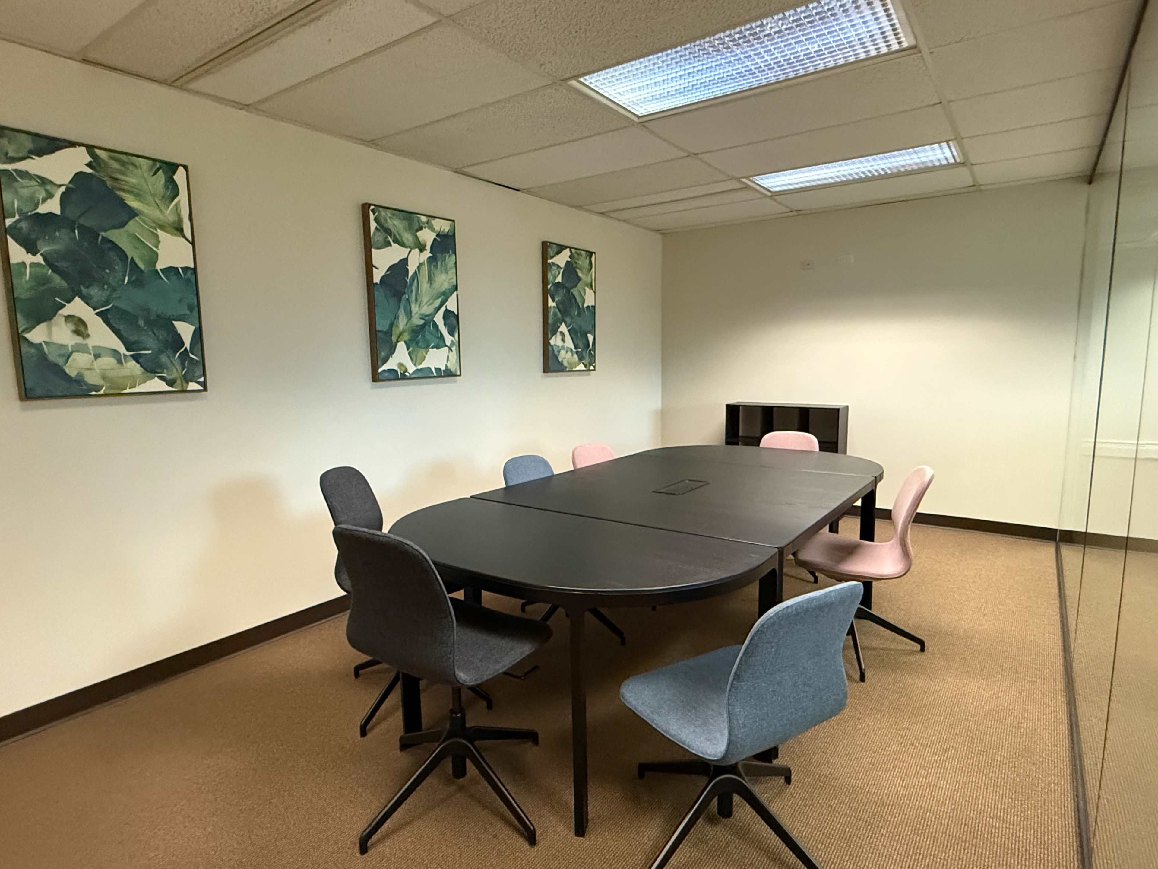 A conference room features a large oval table surrounded by six chairs in various colors, with framed leaf art on the walls.