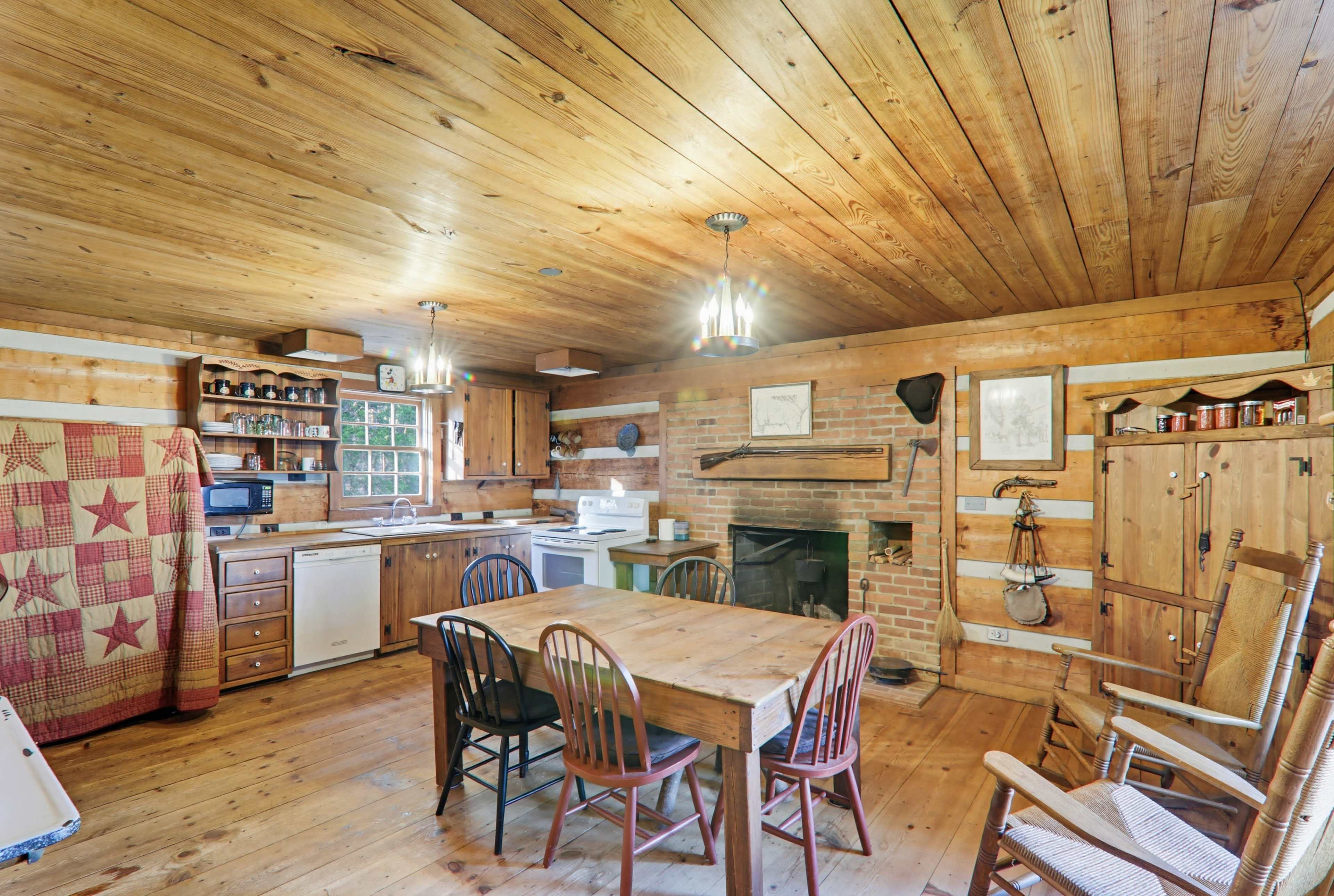 The image shows a rustic kitchen with wooden walls, a large wooden table surrounded by chairs, and a brick fireplace.