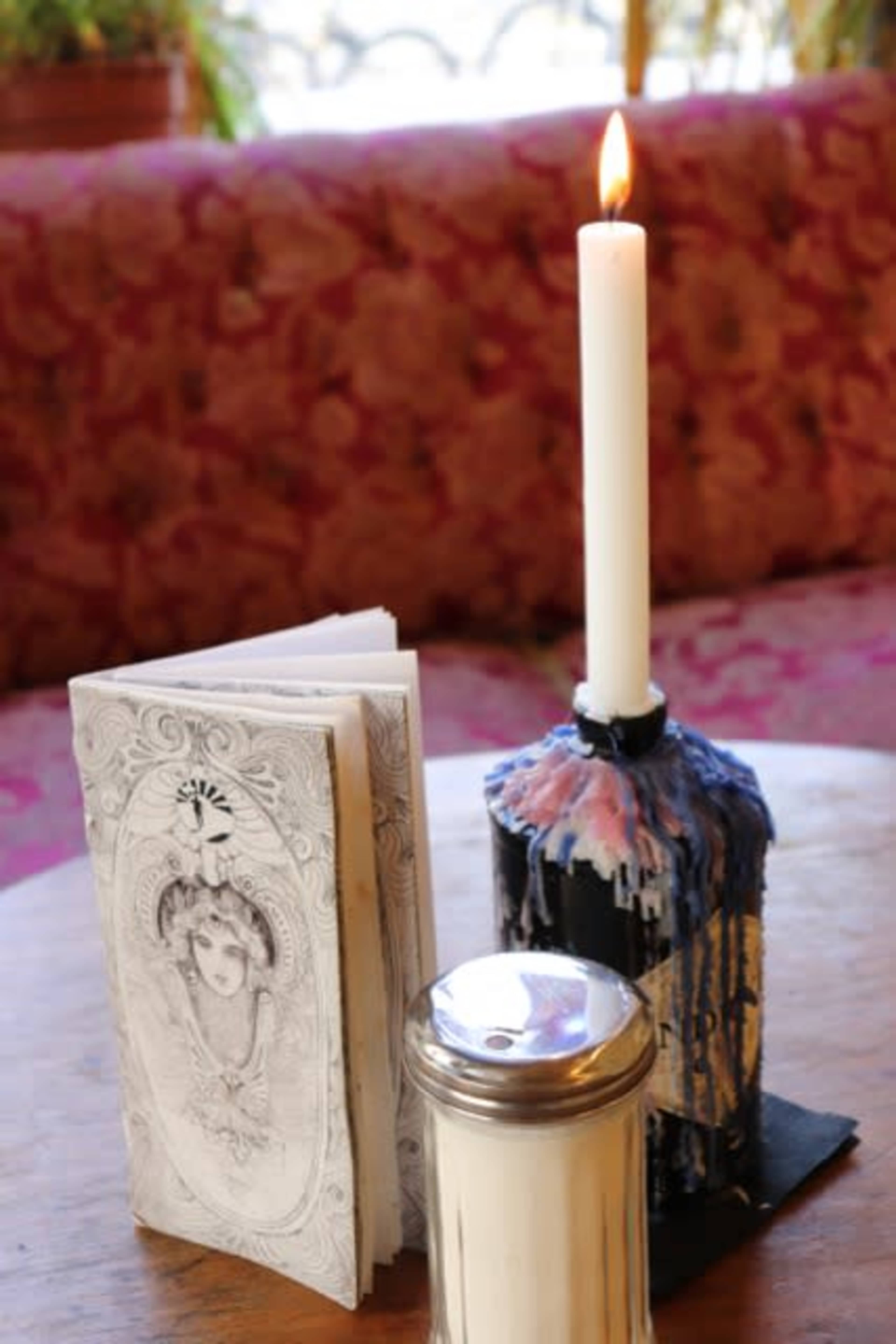 A tall white candle burns in a holder next to a decorative booklet and a metal sugar container on a wooden table.