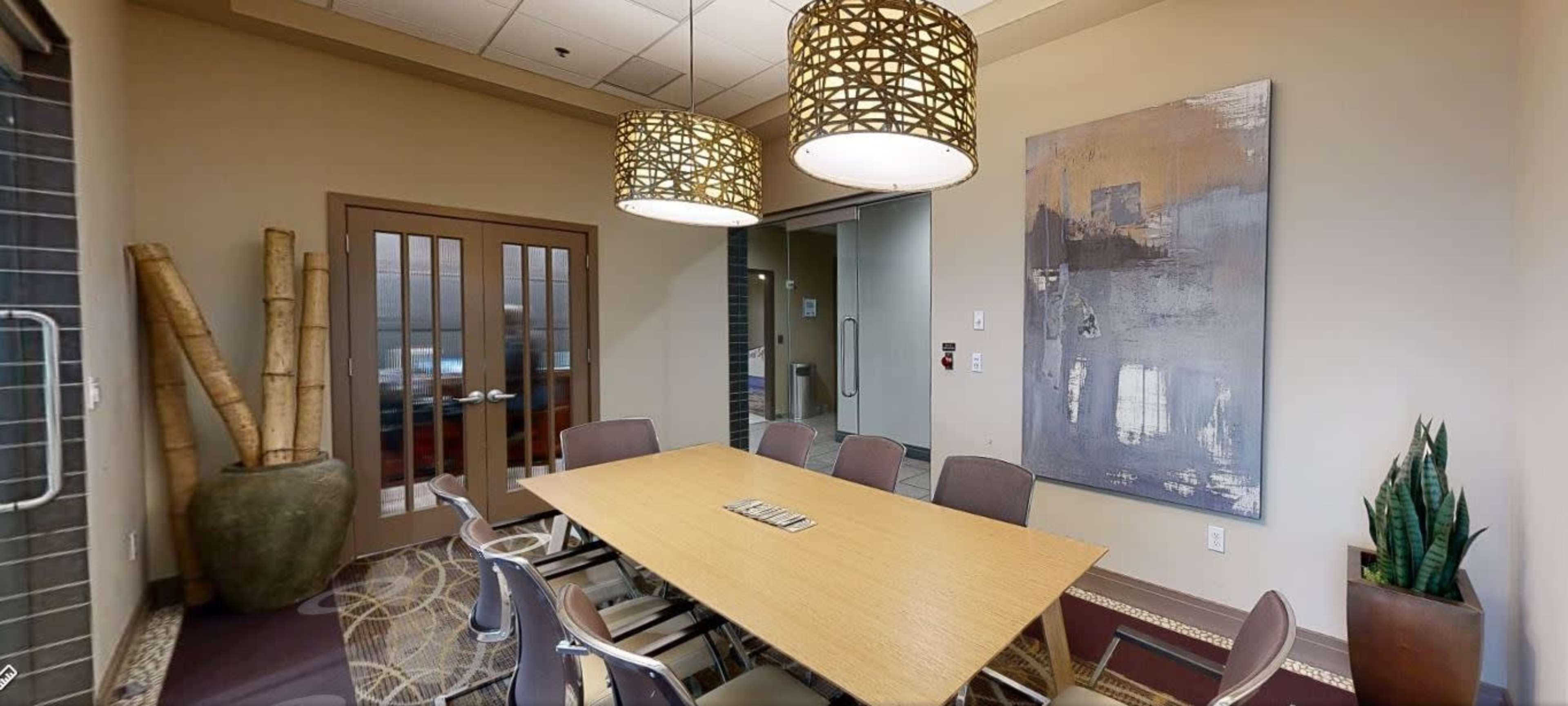 A conference room features a large wooden table surrounded by metal chairs, with two pendant lights overhead and a modern artwork on the wall.