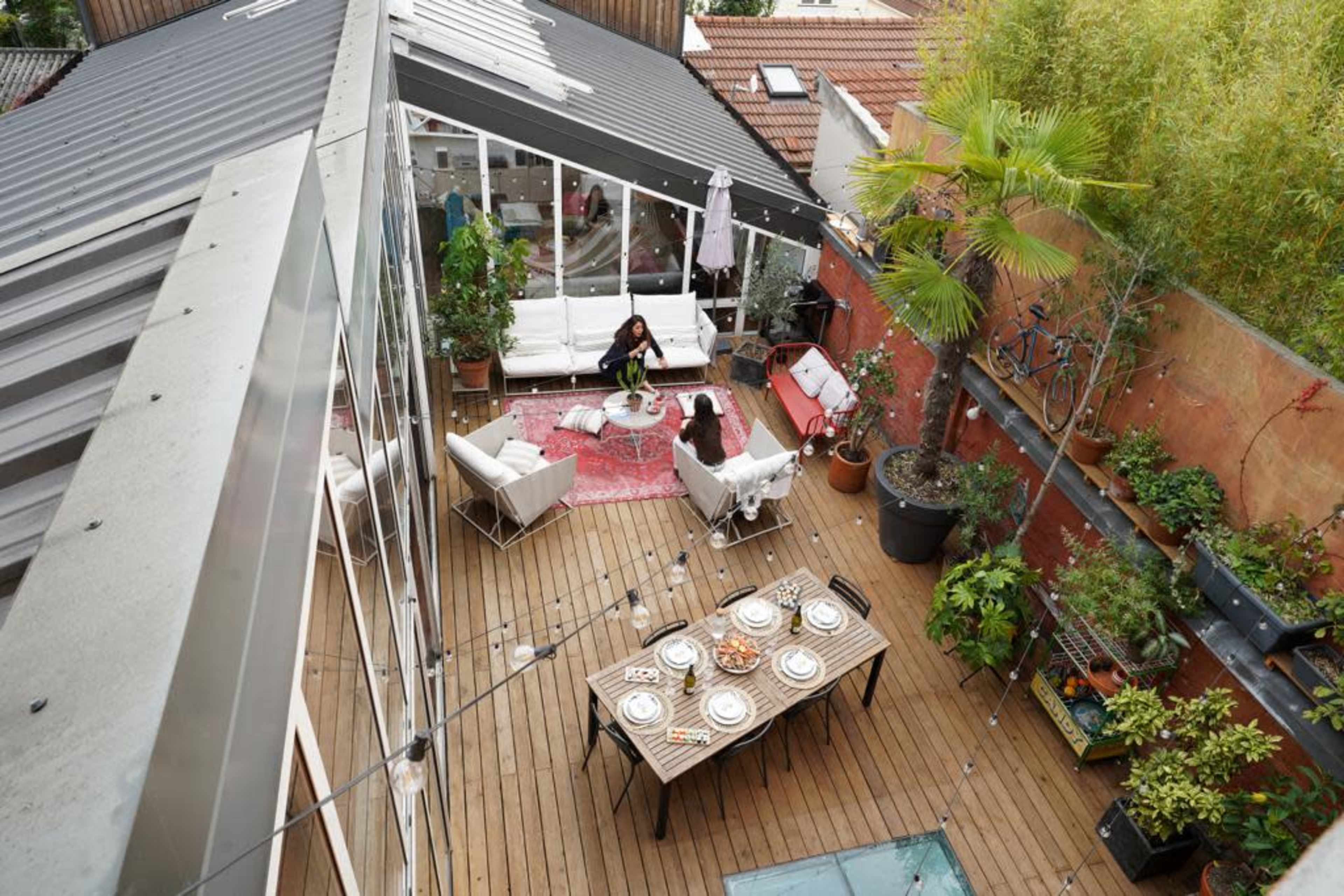 A rooftop patio features a wooden deck with seating arrangements, plants, and a dining table set for a meal.