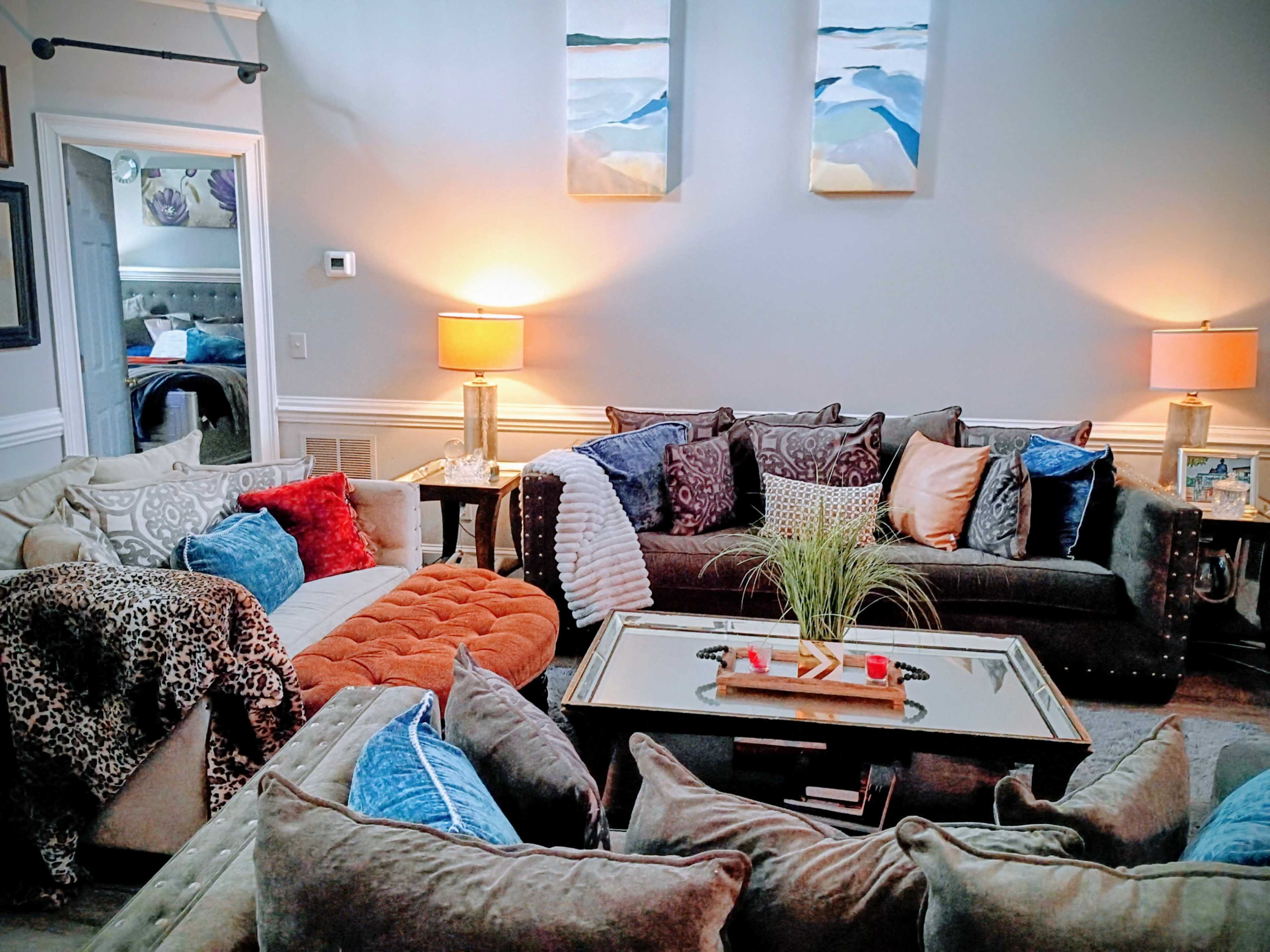 The image shows a cozy living room furnished with various couches and decorative pillows, accompanied by lamps and artwork on the walls.