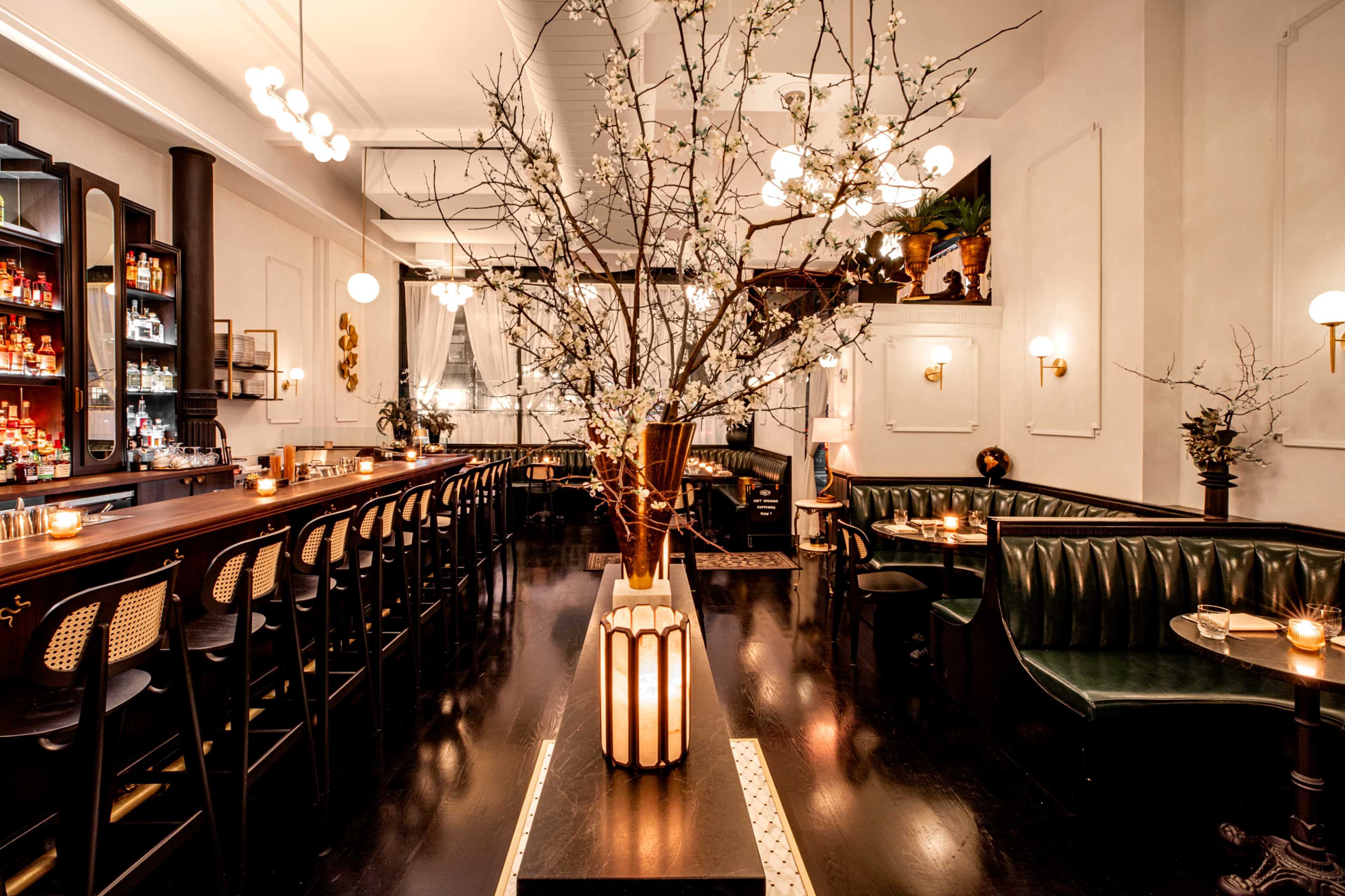 The image shows a stylish bar interior with dark wood furnishings, green leather seating, and decorative branches placed in a vase.