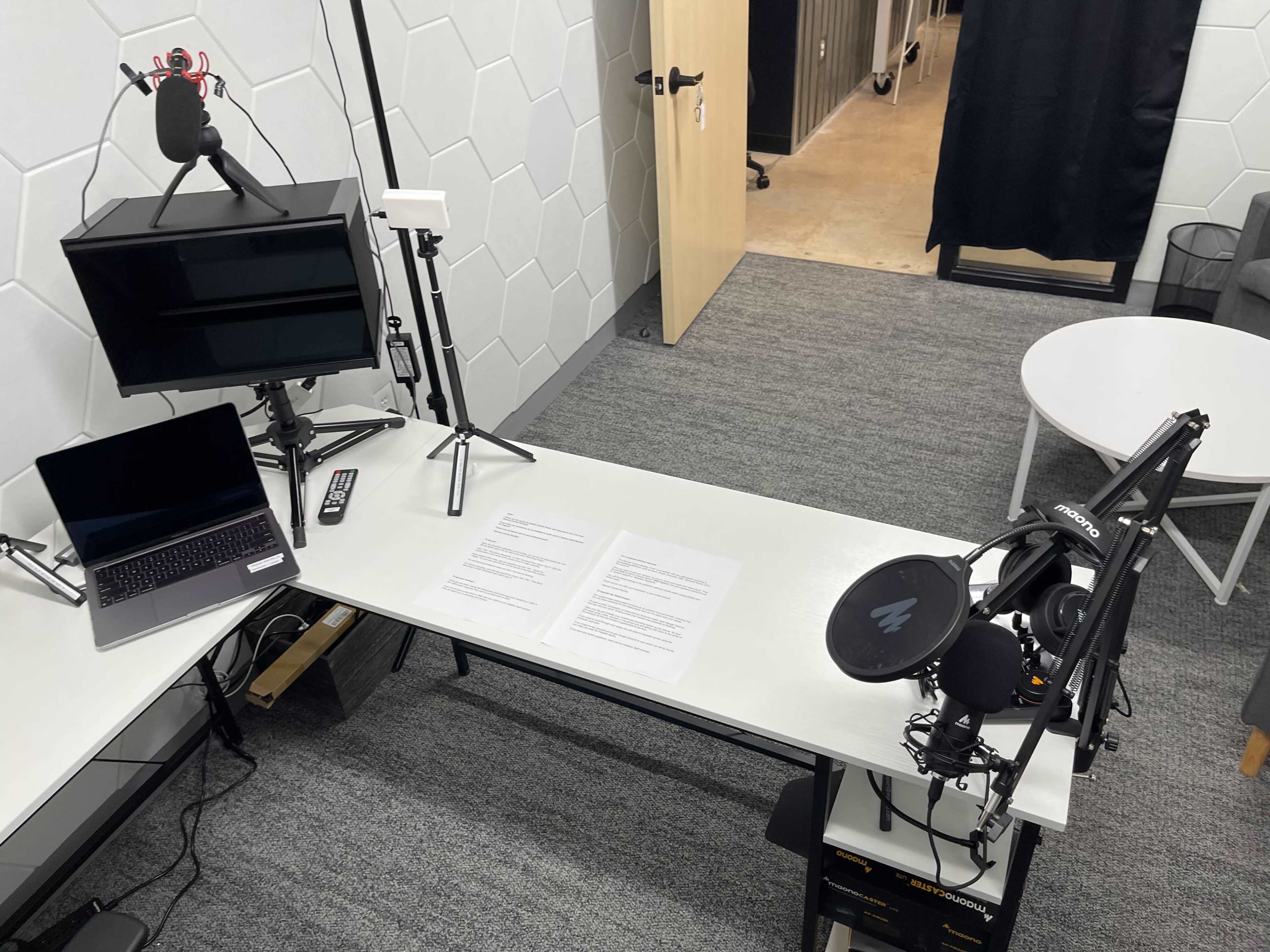 A workspace is set up with a microphone, monitor, laptop, and documents on a white table, surrounded by gray carpeting and walls.