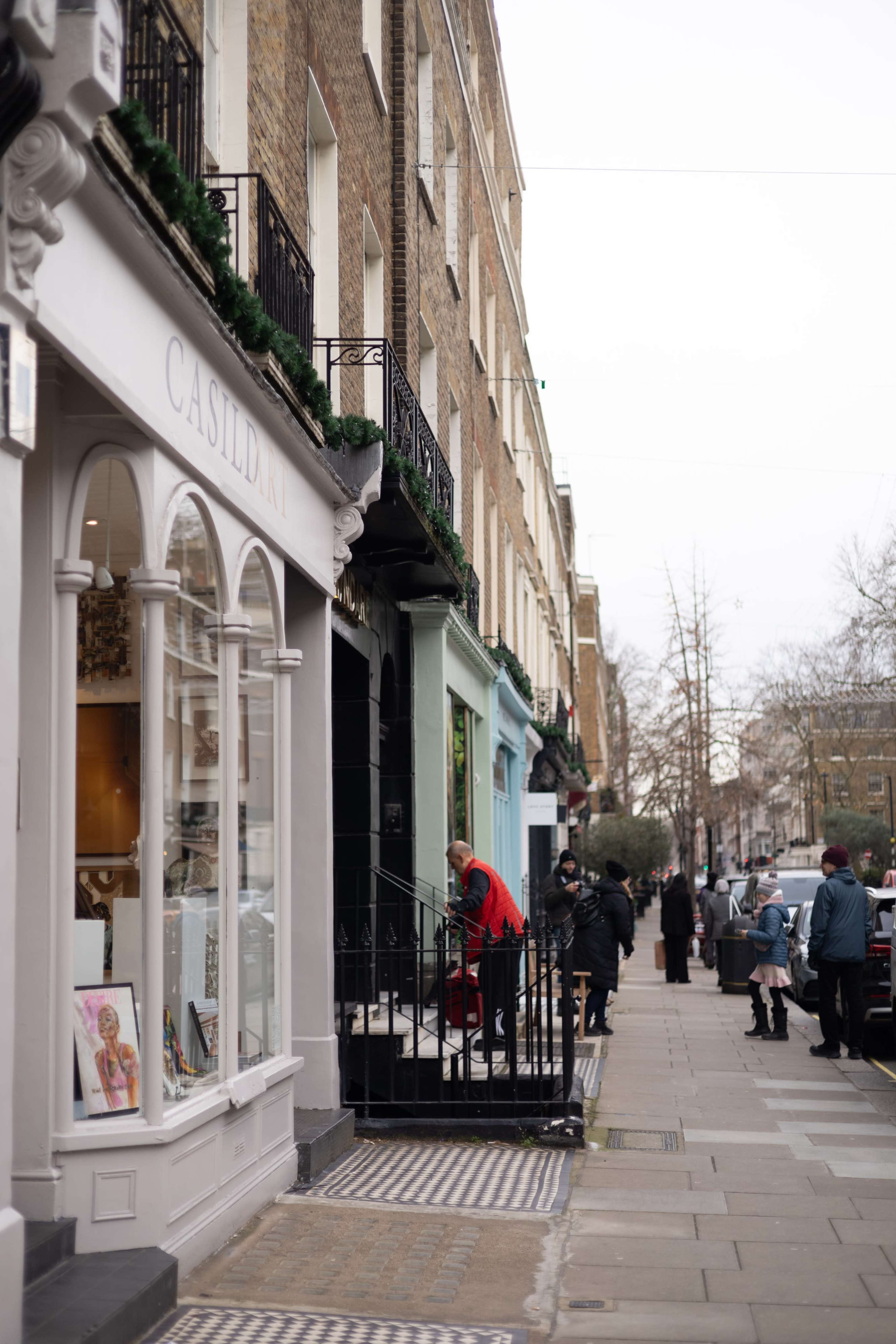 The image shows a street lined with shops and people walking along the sidewalk in a residential area.