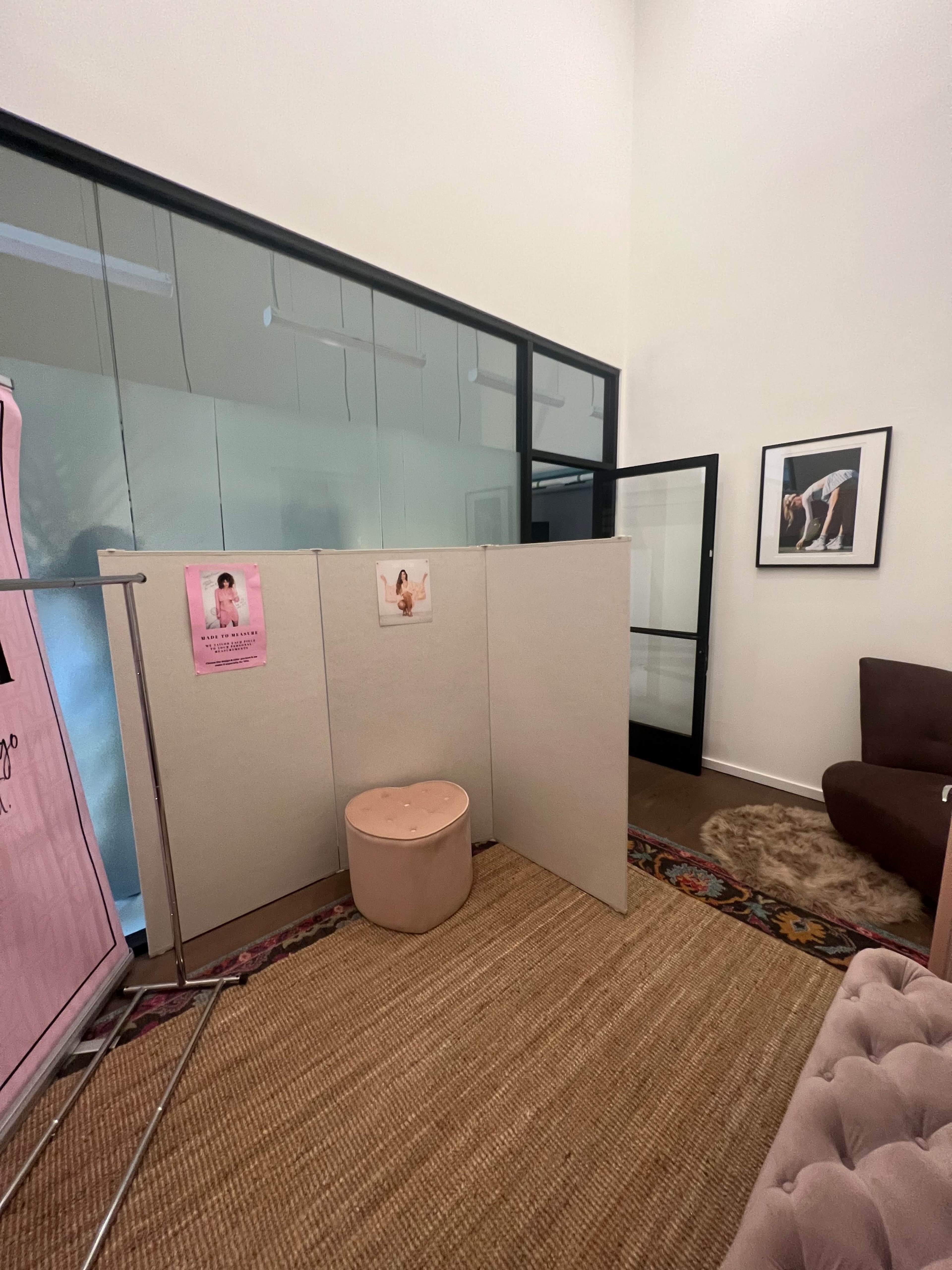 A room with a beige partition, a round stool, and a brown couch, along with posters on the partition and glass walls in the background.