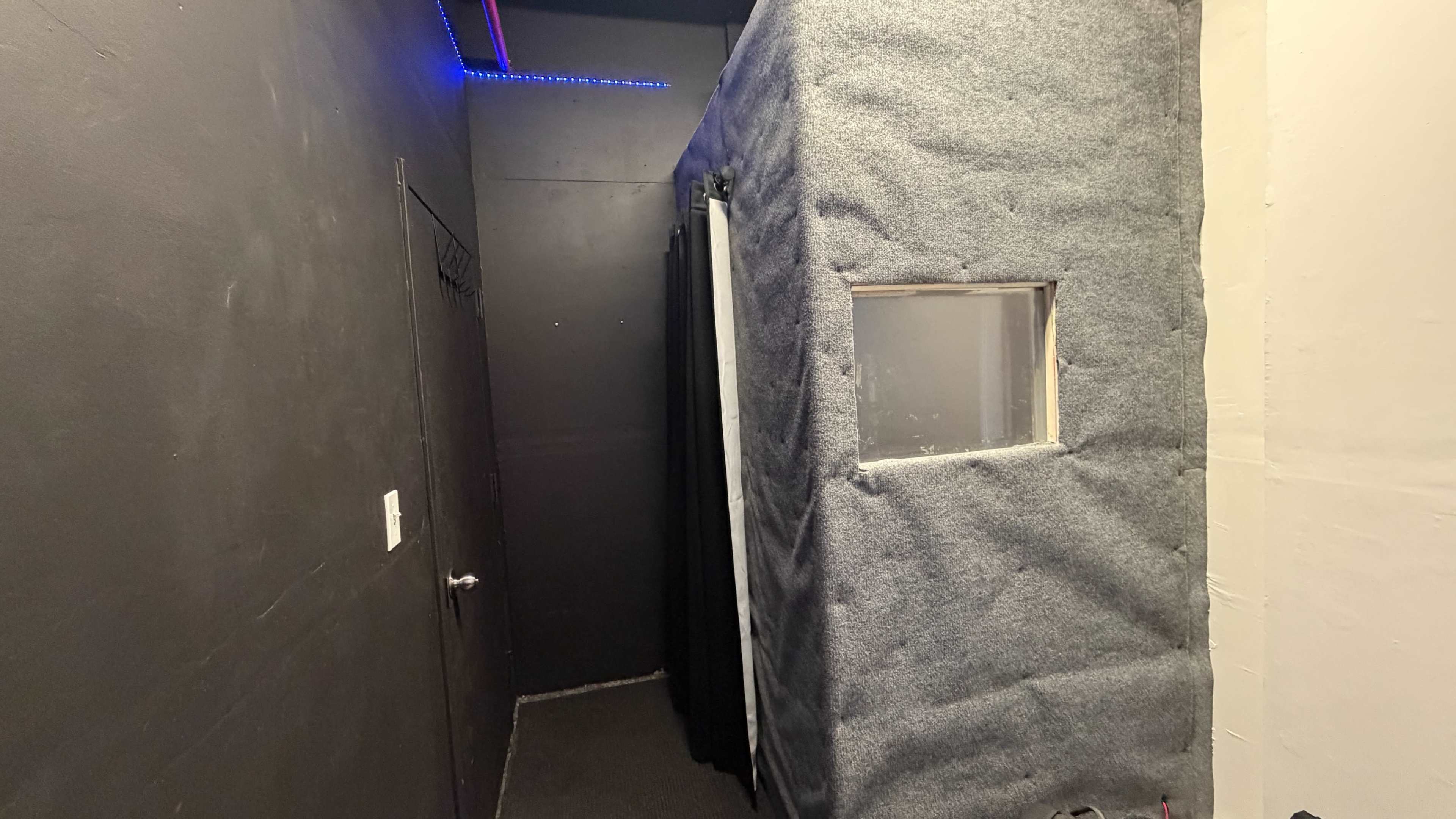 The image shows a narrow hallway leading to a small, enclosed structure covered in gray fabric, with a window and a black curtain nearby.