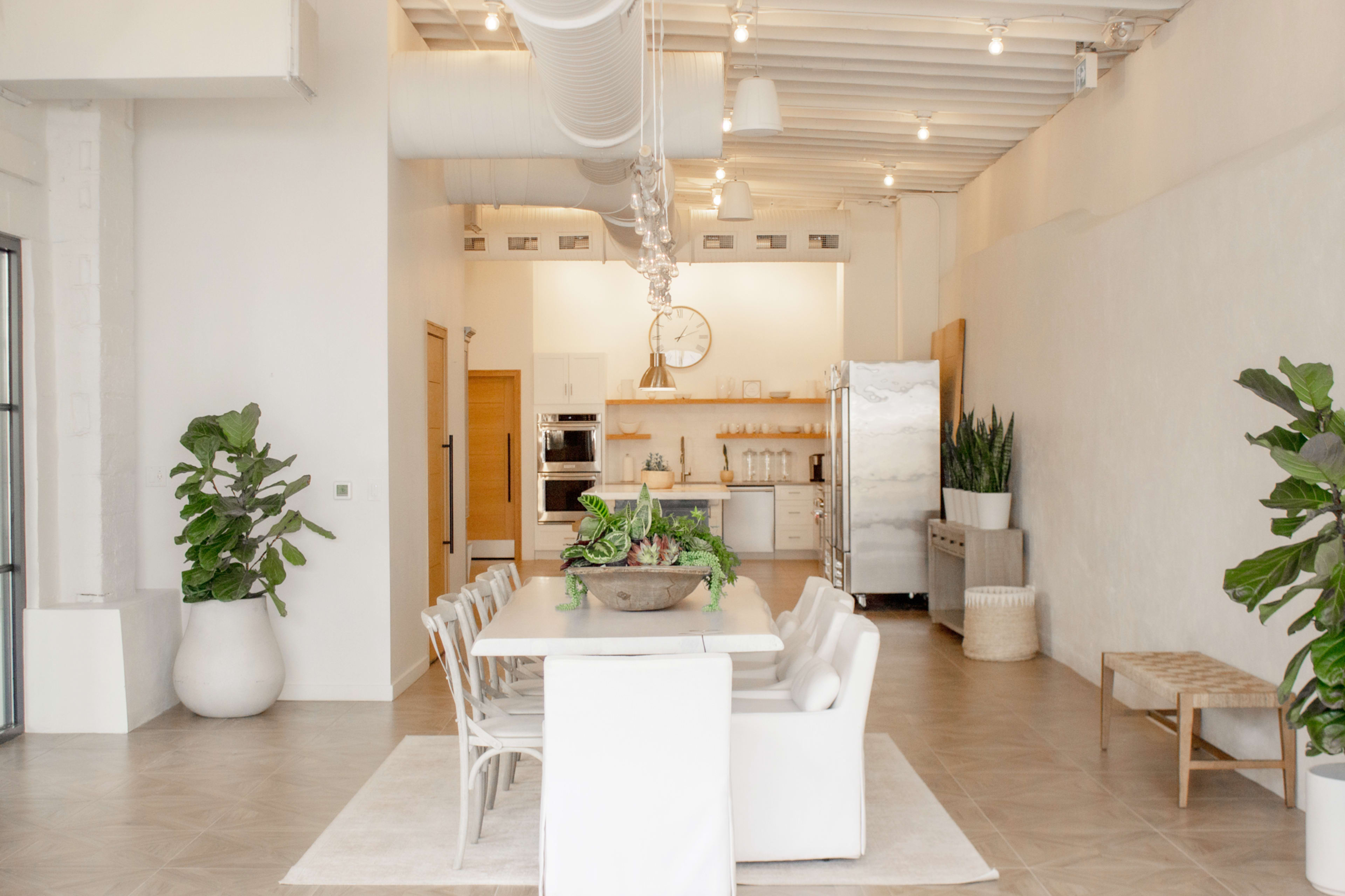 The image shows a modern dining area featuring a rectangular white table surrounded by several chairs, with wall cabinets and a stainless steel refrigerator in the background.