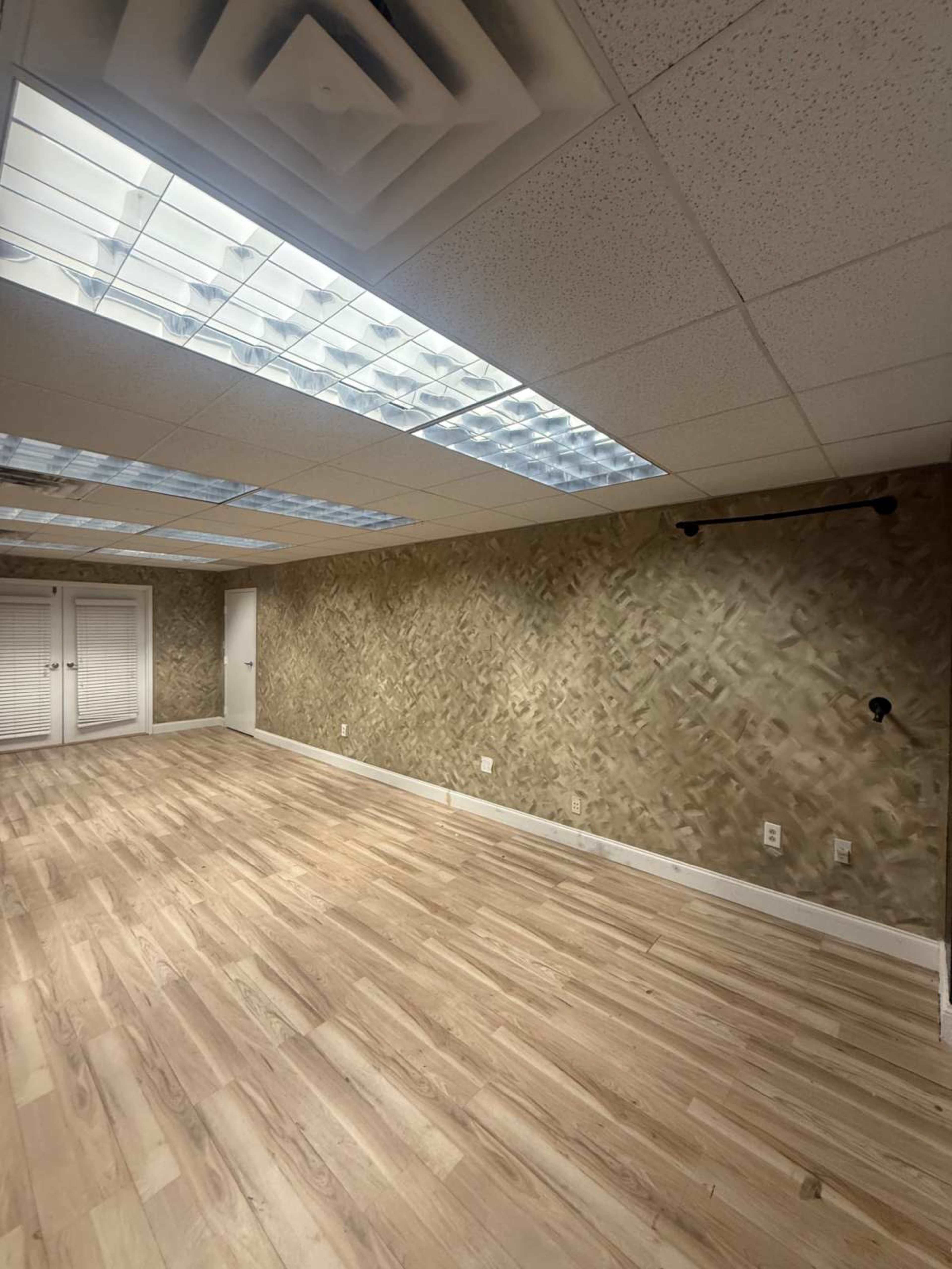 The image shows a spacious, empty room with light-colored wooden flooring and a large skylight providing natural light.