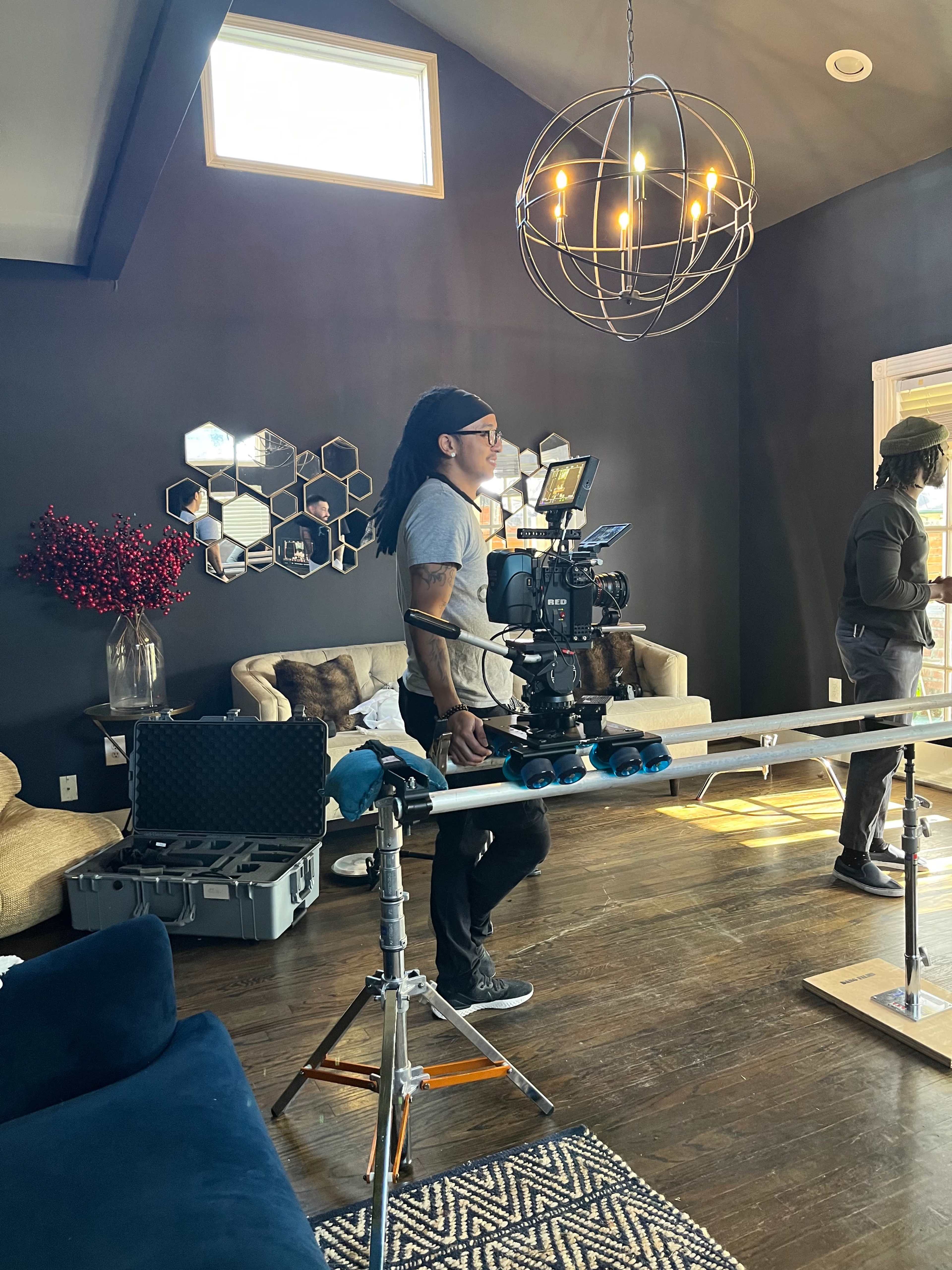 A man operates a camera on a track system in a well-furnished living room with large windows and decorative mirrors.