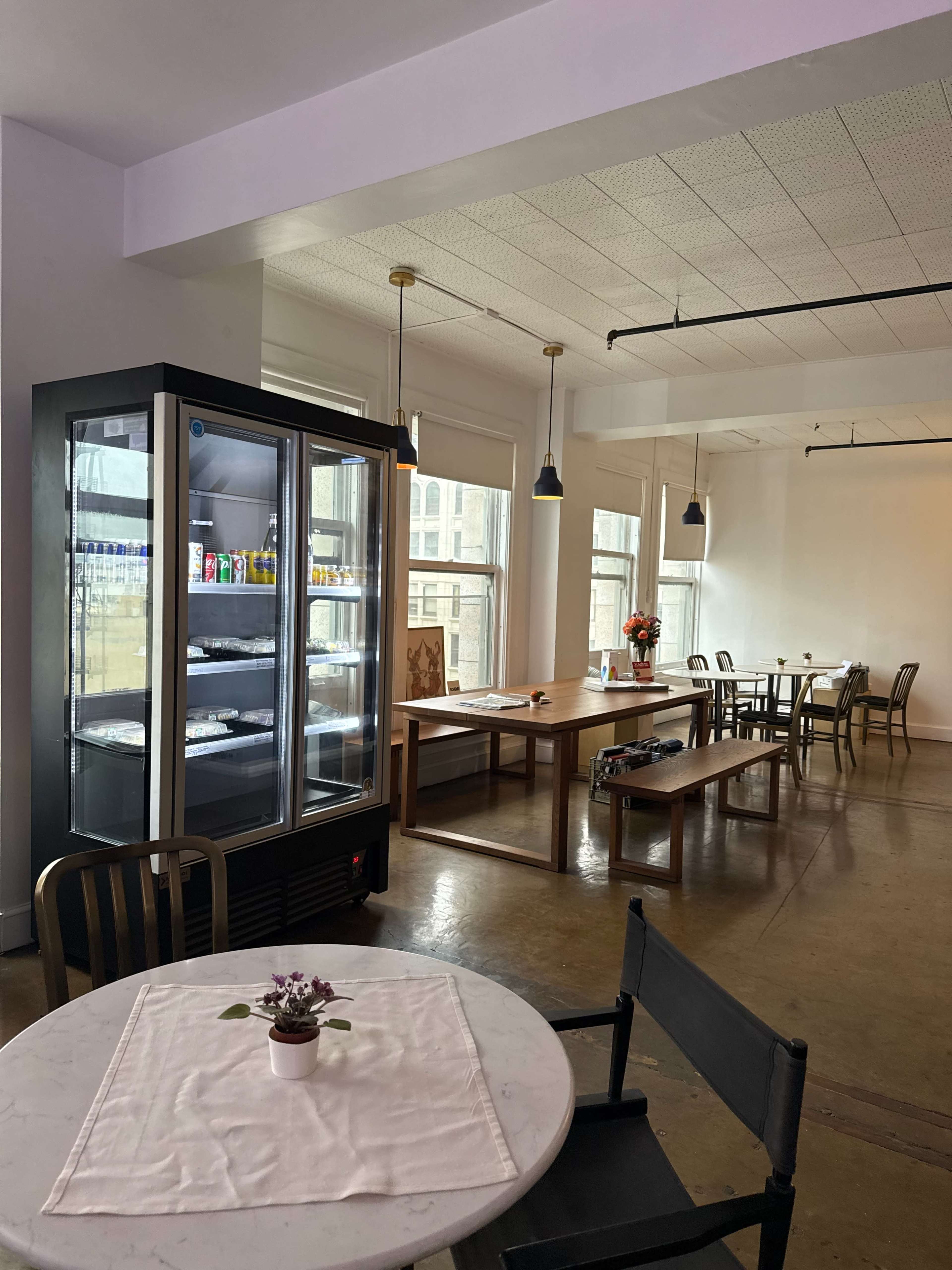 The image shows a bright interior space featuring a refrigerator with beverages, a simple table setup, and large windows allowing natural light to enter.
