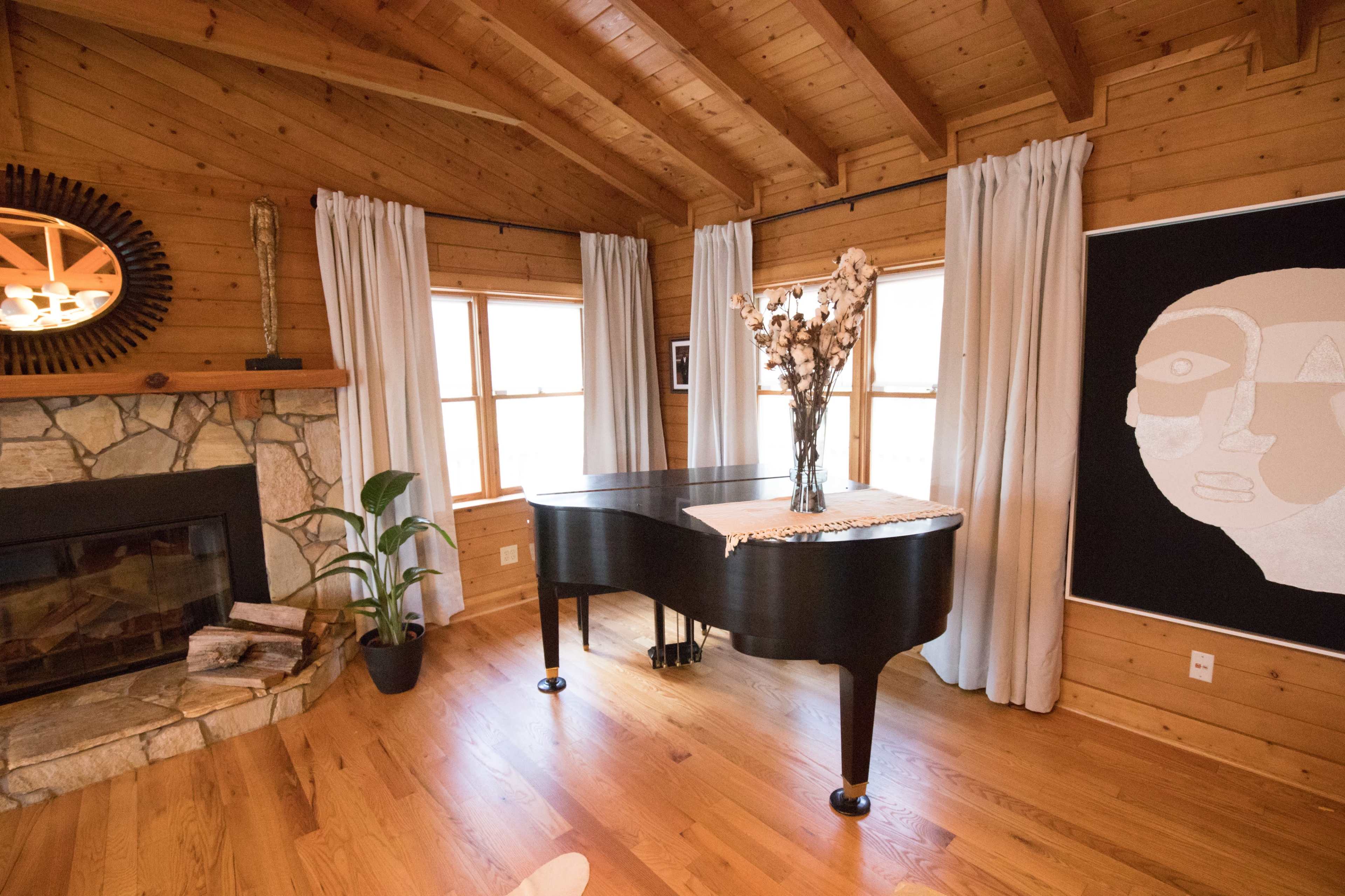A grand piano sits in a wooden room beside a stone fireplace, with large windows framed by curtains and a decorative wall art nearby.