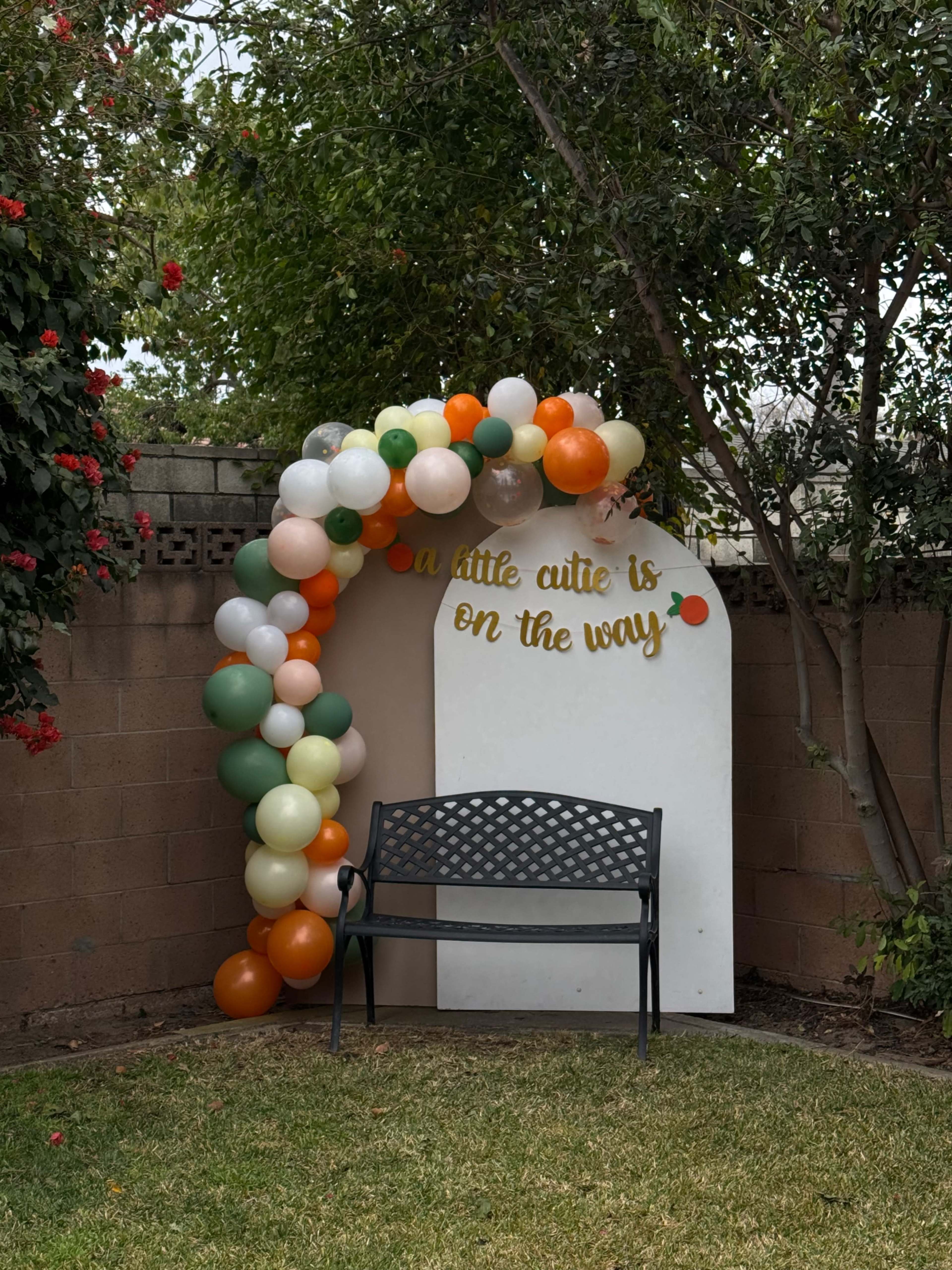 A white arch decorated with balloons in various colors stands next to a bench in a garden setting.