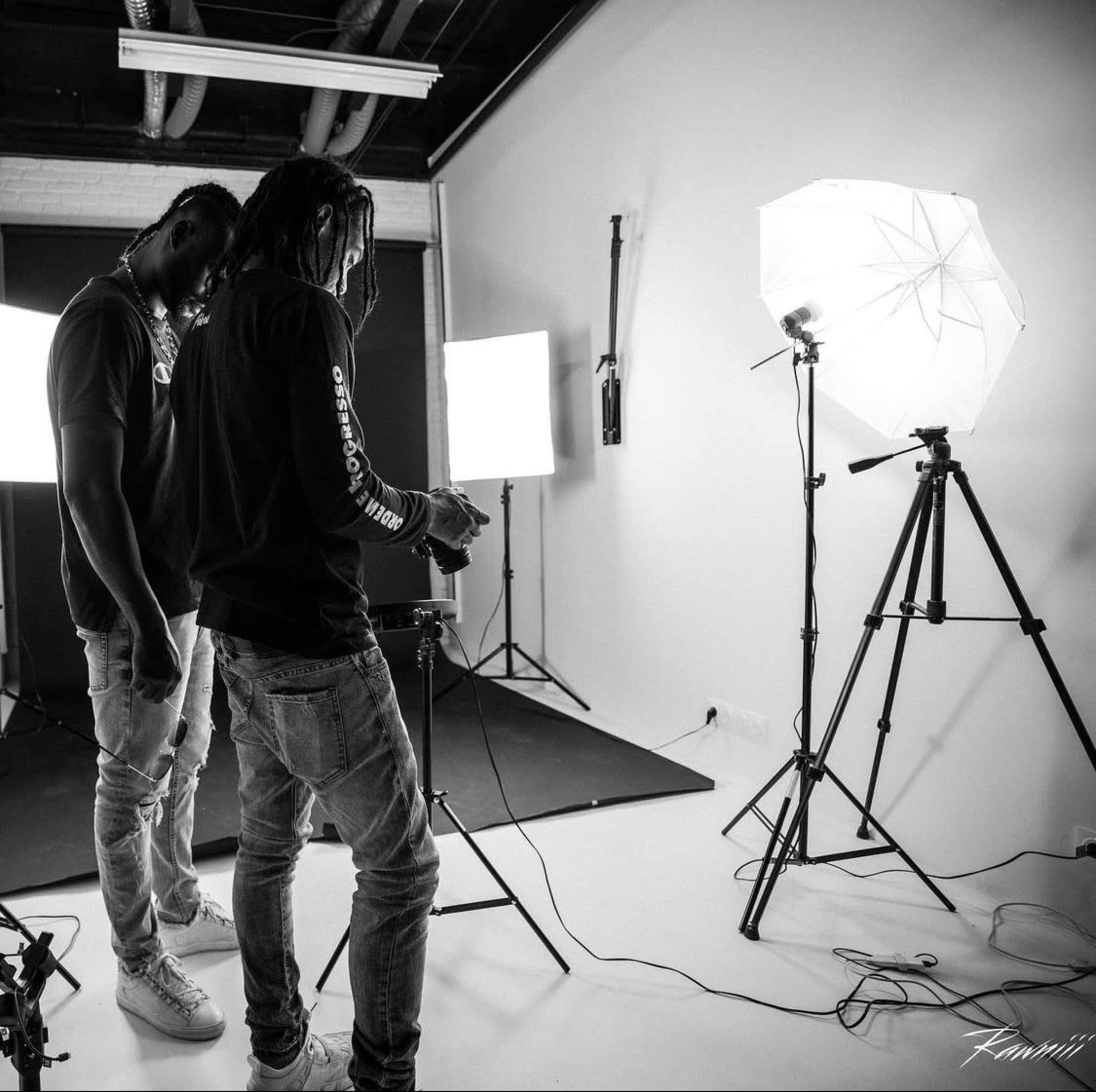 Two individuals set up photography equipment in a studio with softbox lights and a camera on a tripod.