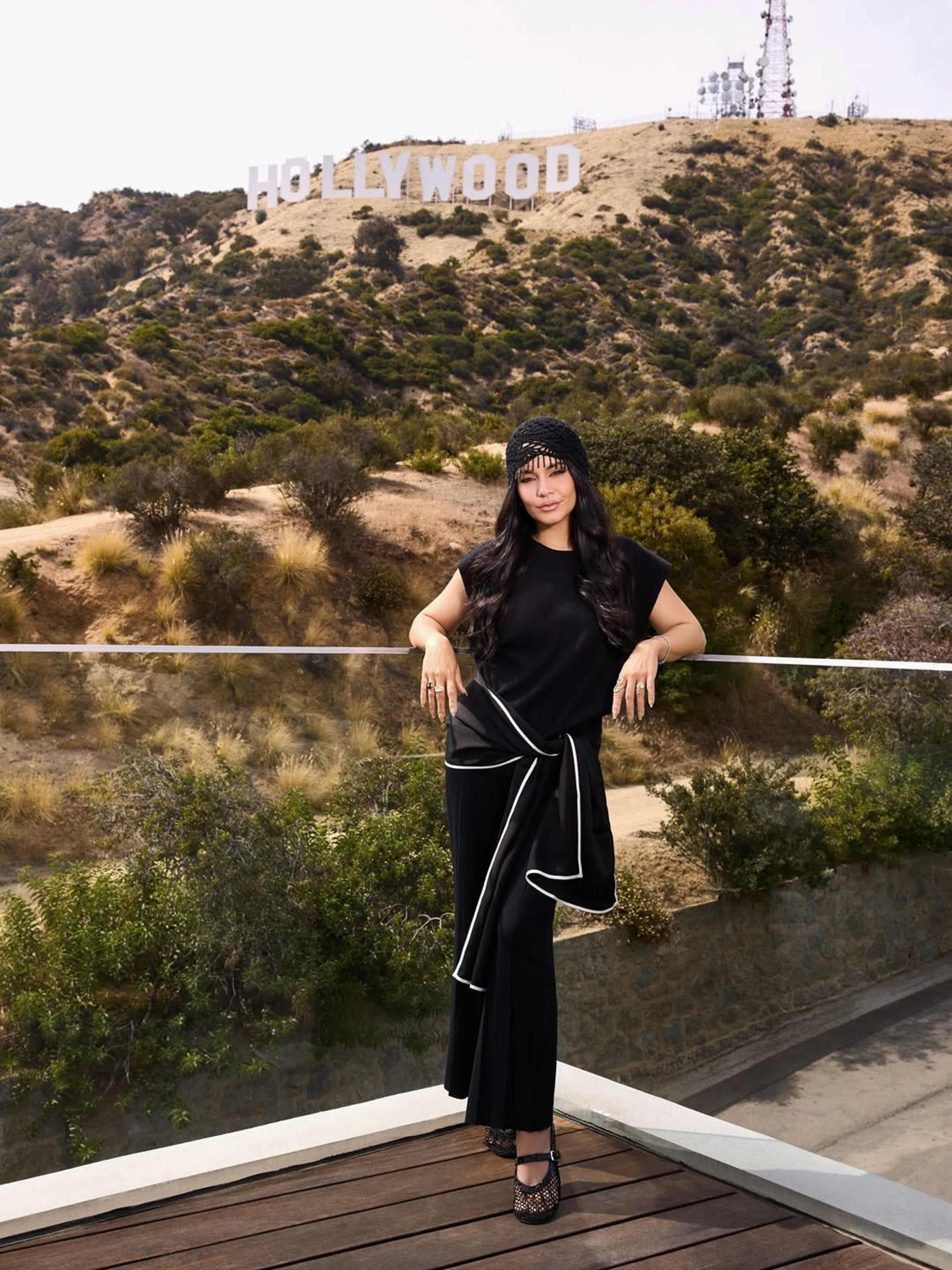 A person poses in a black outfit on a balcony with the Hollywood sign visible in the background.